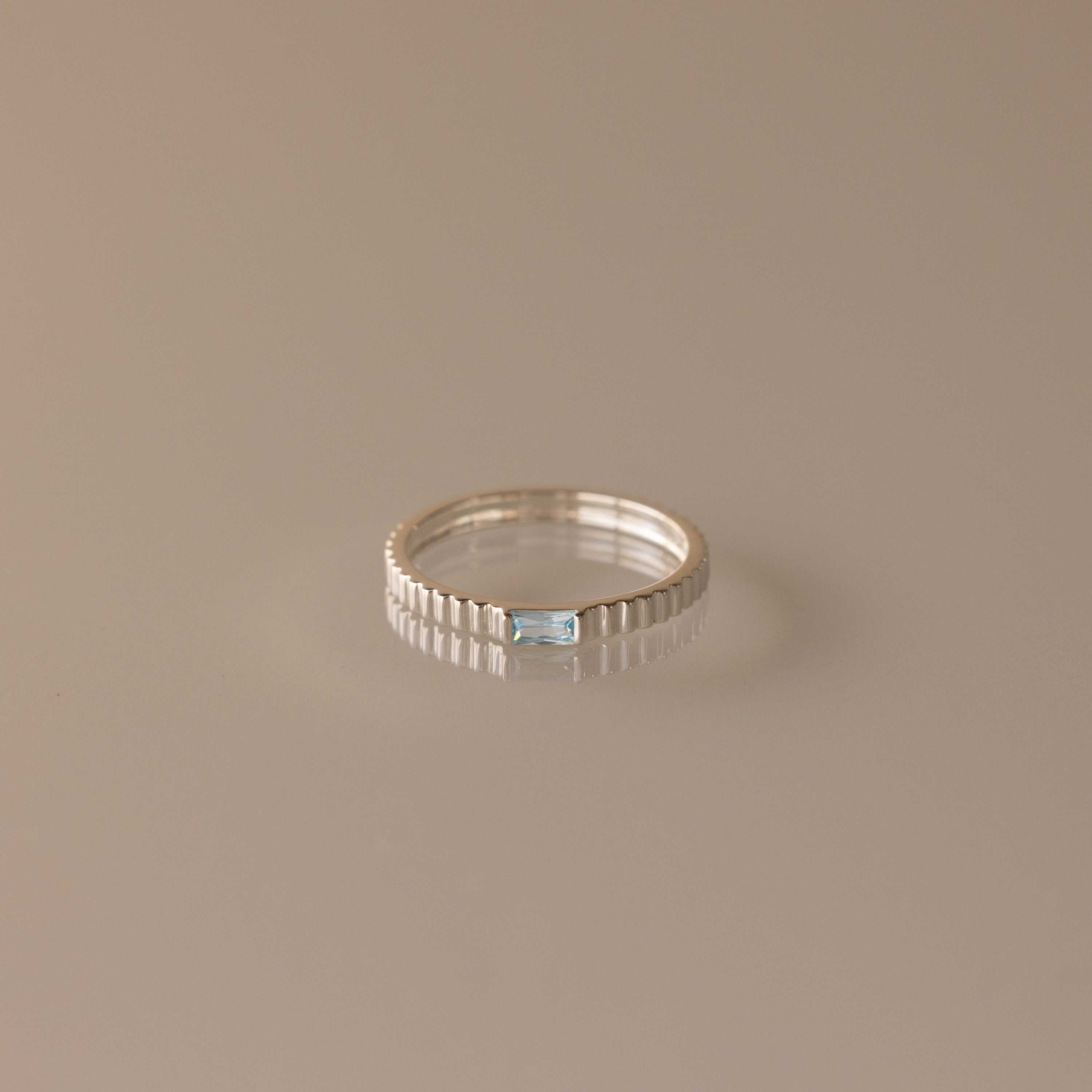 The Dainty Ribbed Aquamarine Ring, featuring a rectangular blue gemstone, is shown on a smooth beige surface.