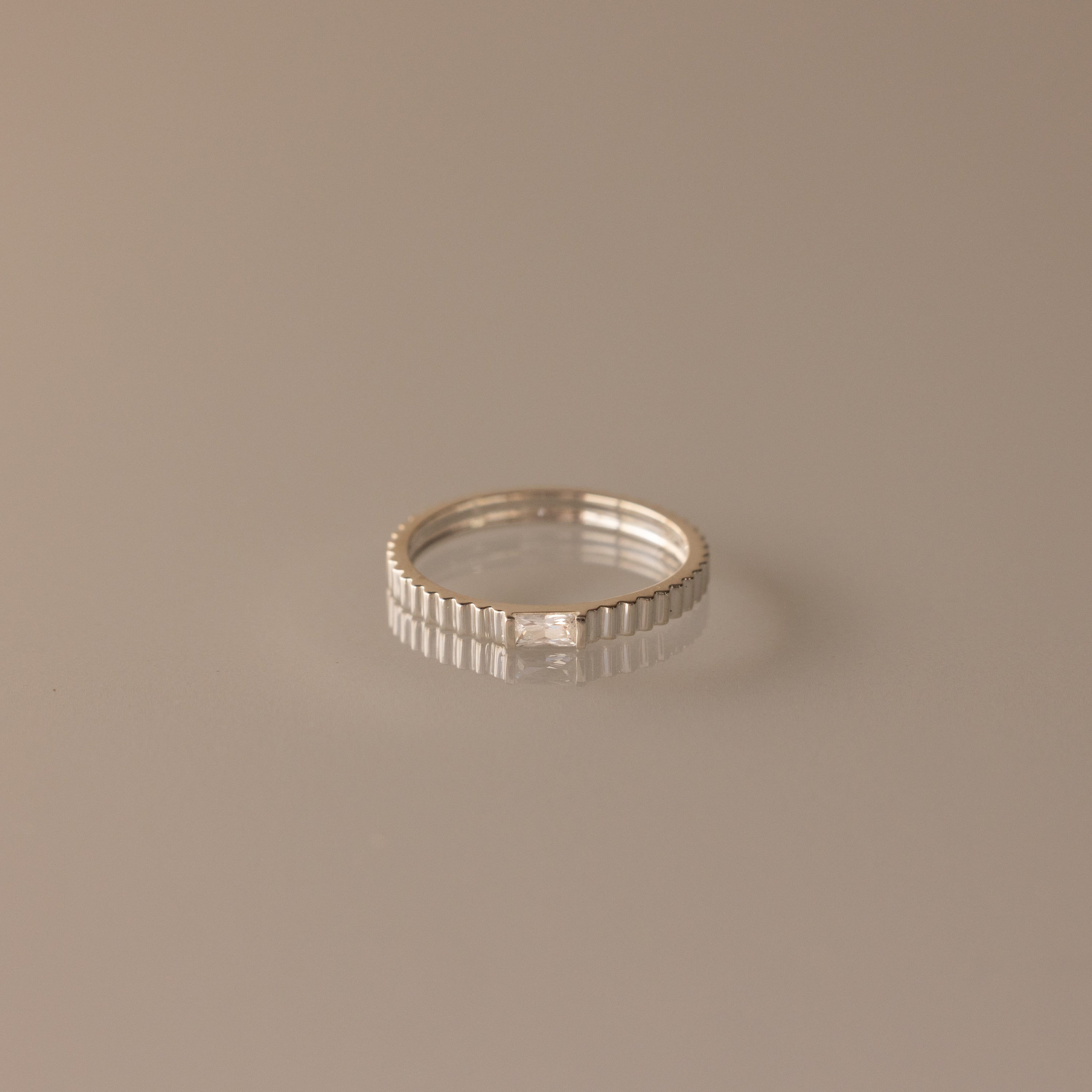 The Dainty Ribbed Diamond Ring features a slender silver ridged band with a small diamond, ideal for stacking or as a subtle birthstone ring on a neutral background.