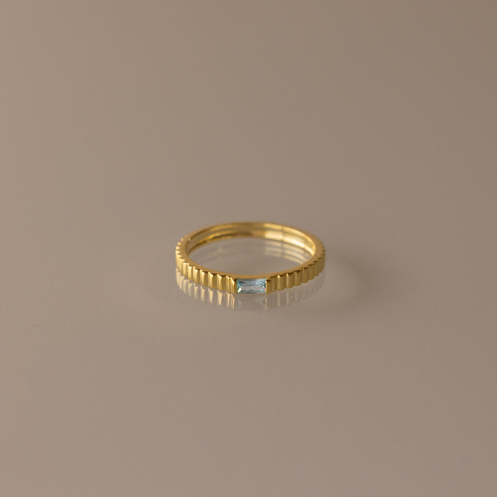 The Dainty Ribbed Aquamarine Ring features a gold band with a rectangular aquamarine gemstone at its center, shown against a neutral background.