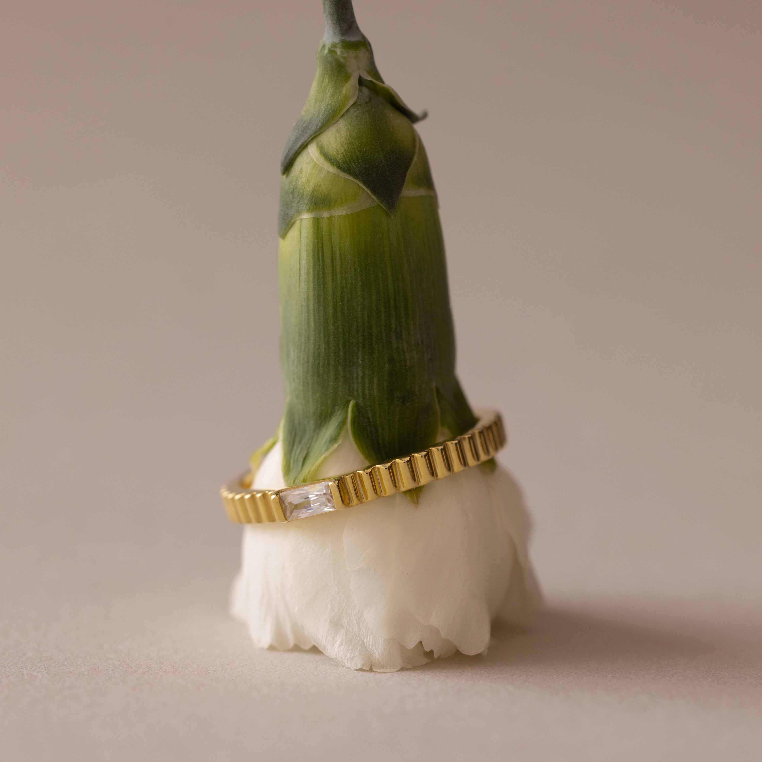 The Dainty Ribbed Alexandrite Ring, featuring a rectangular gemstone, rests on a partially bloomed white flower bud against a beige background.