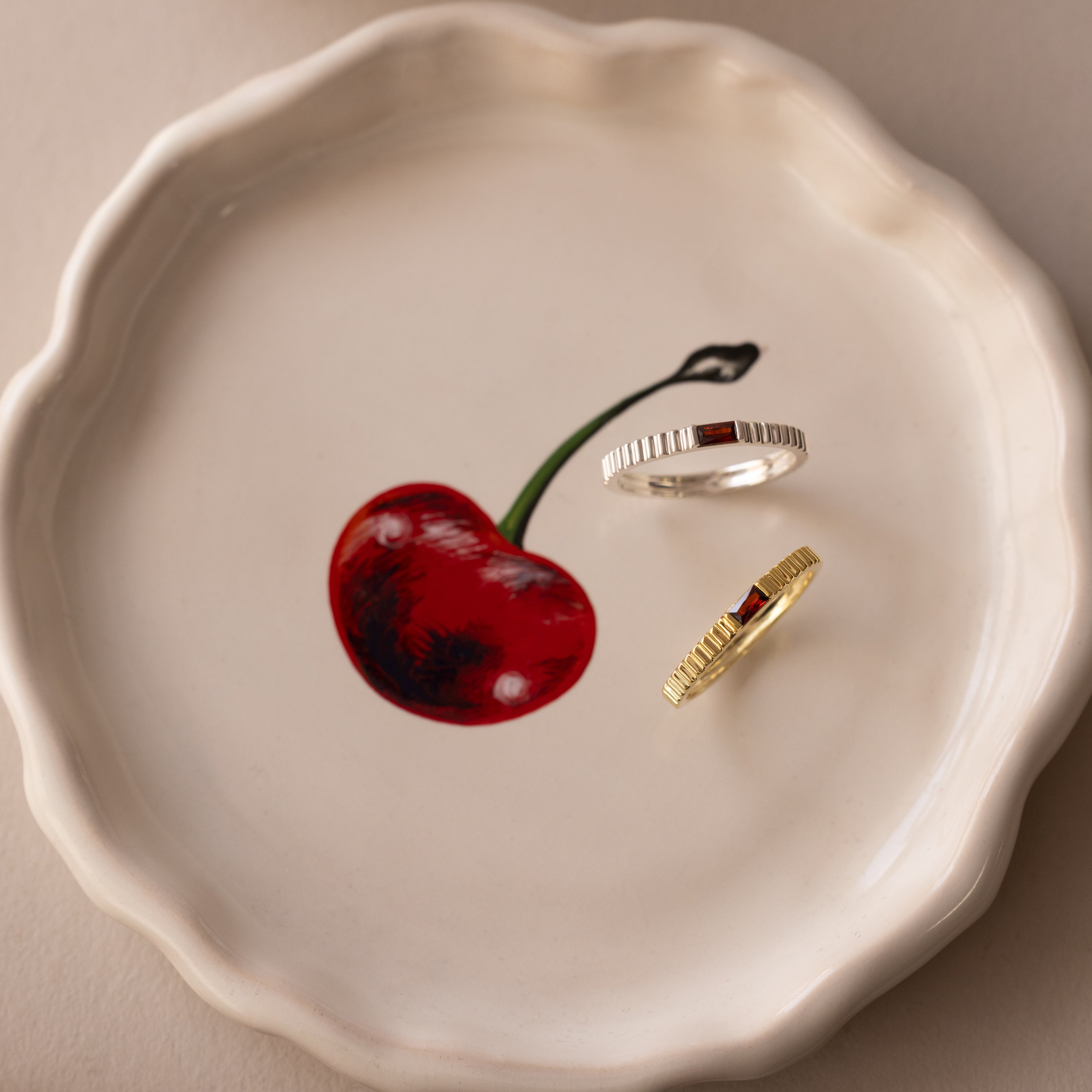 A Dainty Ribbed Birthstone Ring in silver and another in gold rest stylishly on a scalloped white dish with a painted red cherry—perfect for stacking or wearing solo.