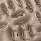 The Dainty Ribbed Ruby Ring features a textured, ribbed band and a vibrant ruby gemstone, displayed on a beige surface—perfect for stacking with other rings.