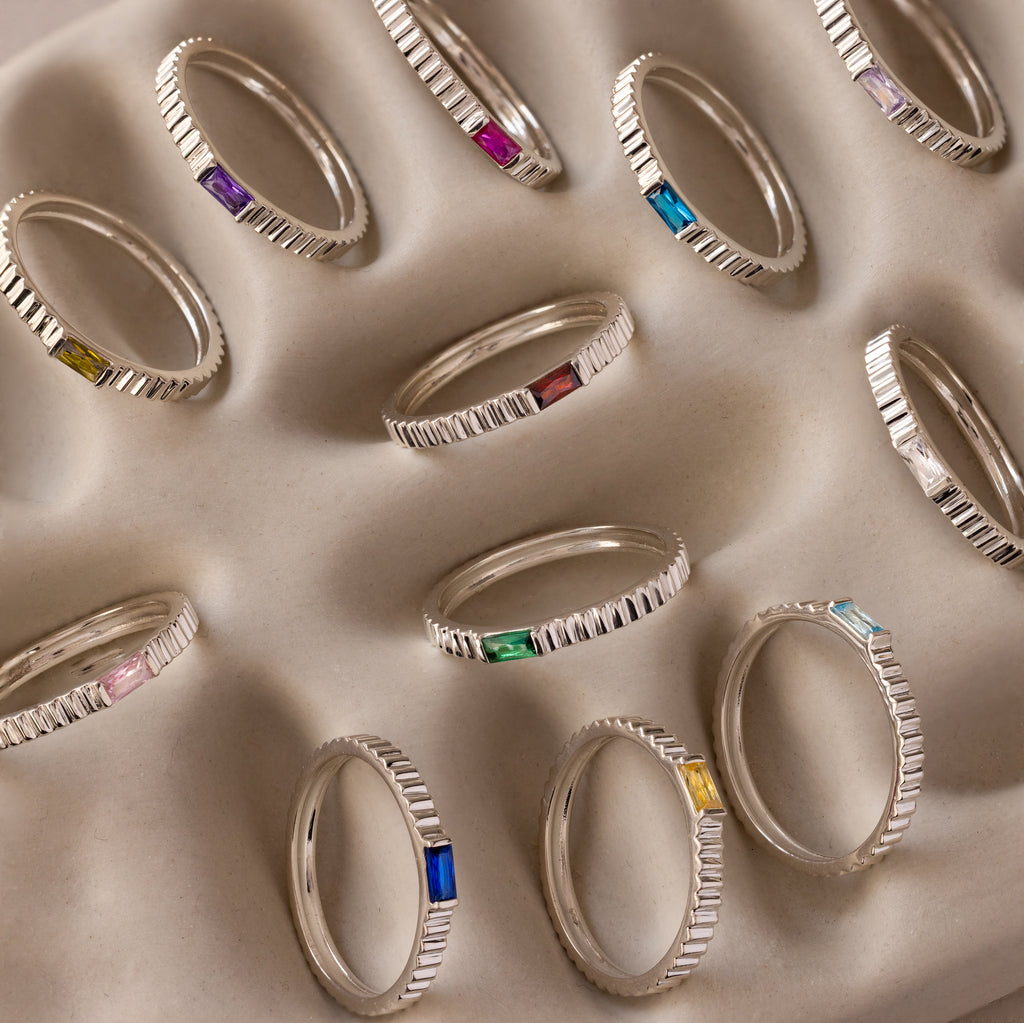 The Dainty Ribbed Topaz Ring, featuring a vibrant gemstone, is perfect for stacking or wearing alone as a subtle birthstone accent, shown neatly arranged on a textured beige surface.