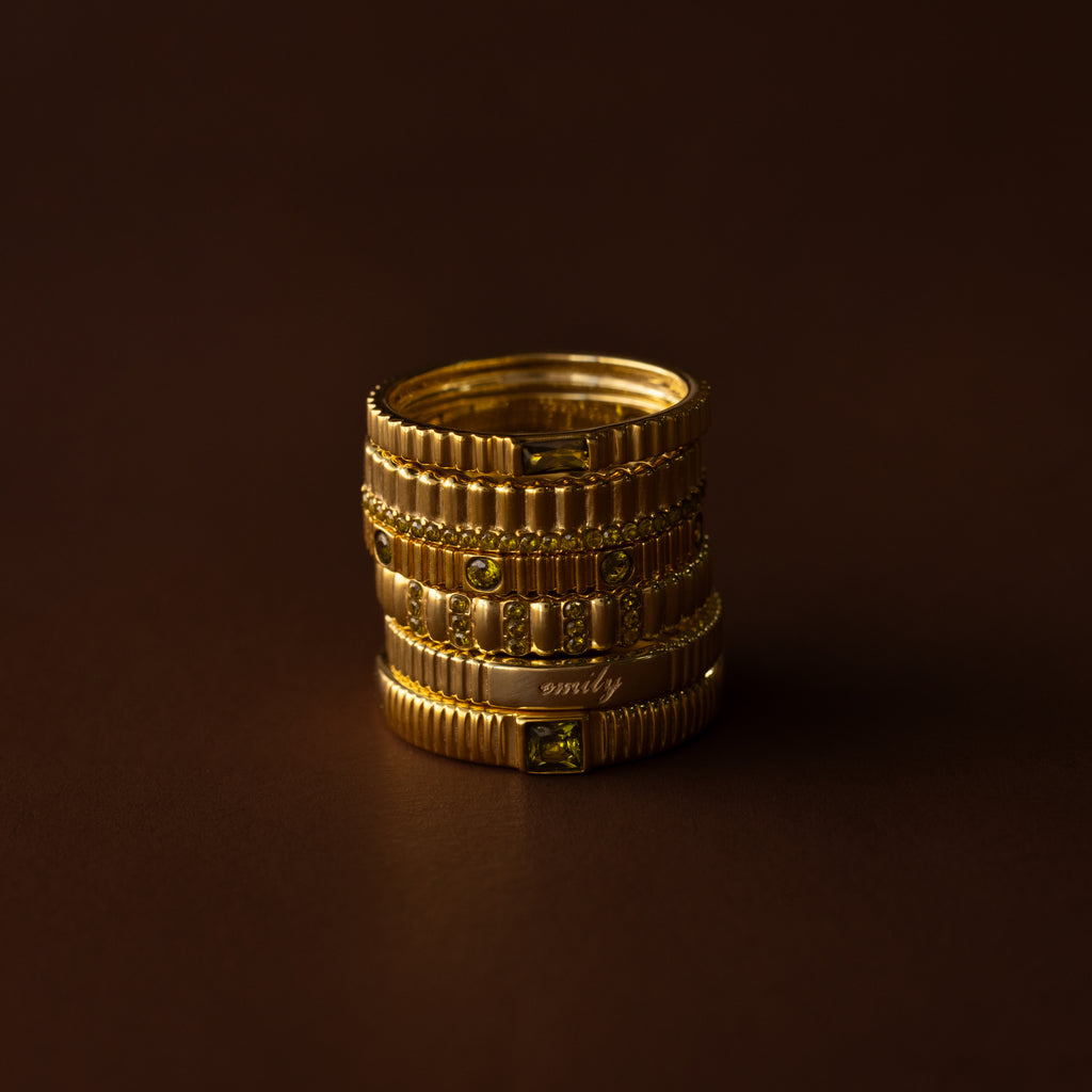 A Dainty Ribbed Birthstone Ring with textured gold bands and green gemstones is stacked together, displayed on a dark brown background.