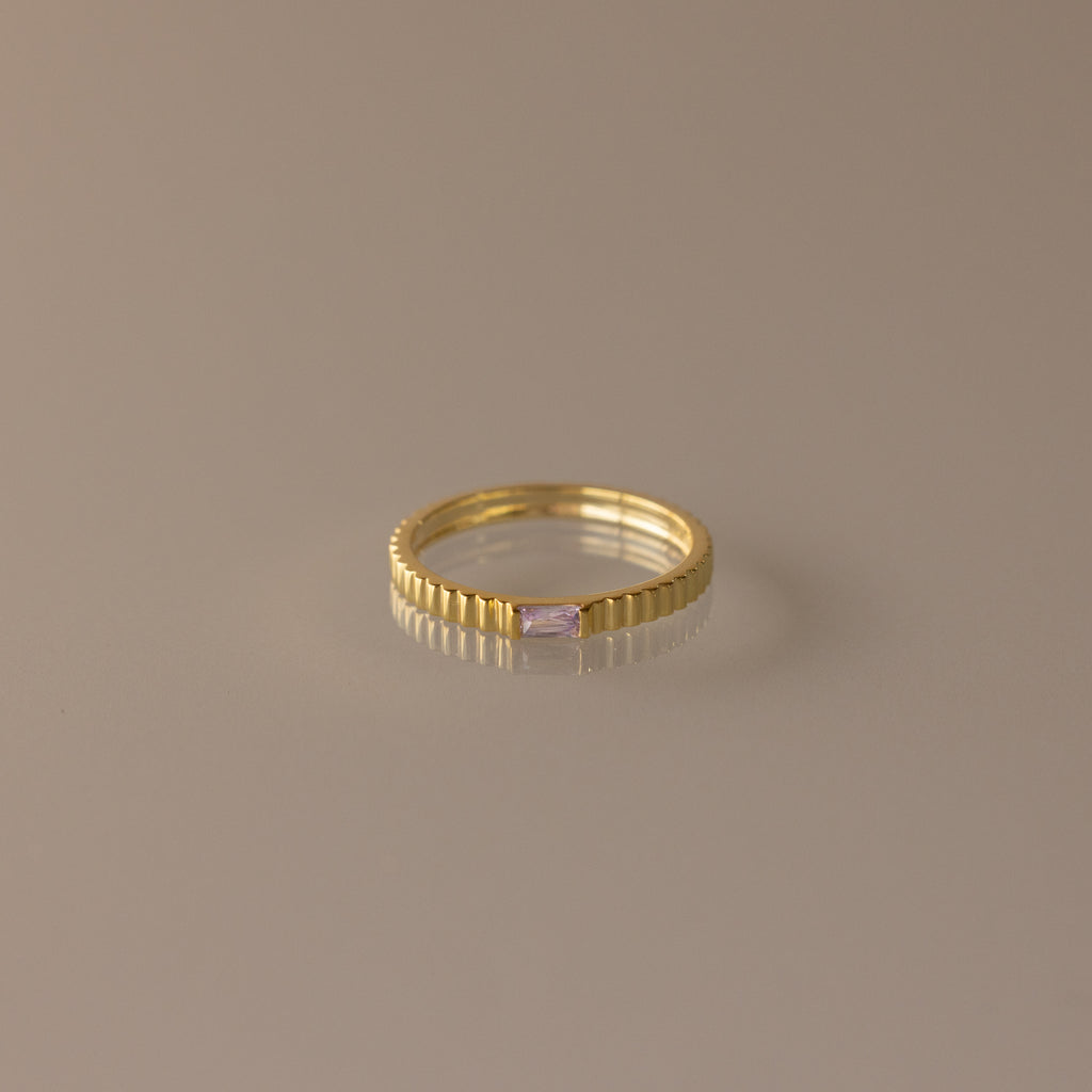 The Dainty Ribbed Alexandrite Ring features a gold ridged band and a small rectangular pale pink gemstone, ideal as a delicate ring or unique birthstone piece. Displayed on a plain surface.