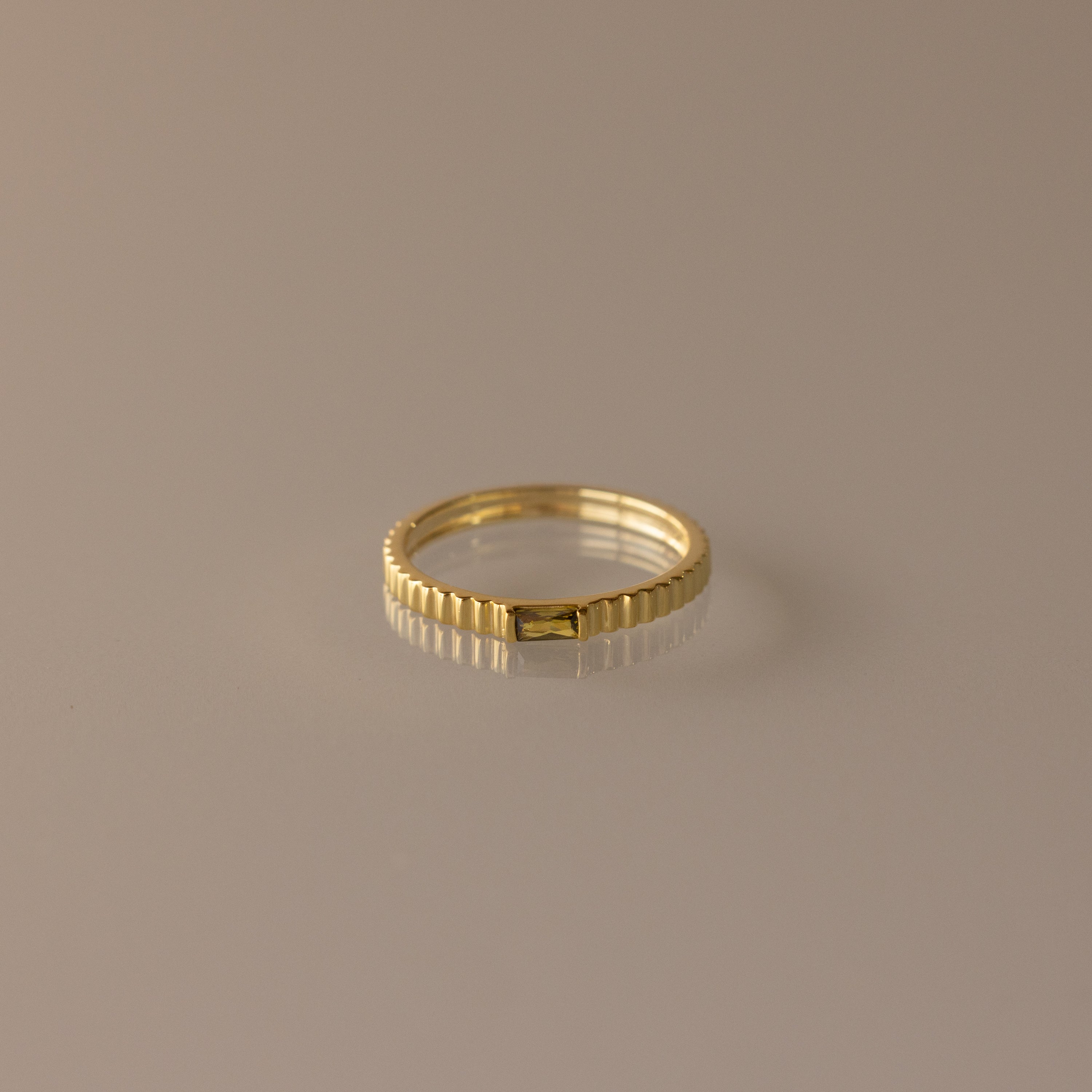 The Dainty Ribbed Peridot Ring features a gold, ribbed design and a minimalist look, displayed on a neutral background—perfect for stacking and adding elegant style to any outfit.