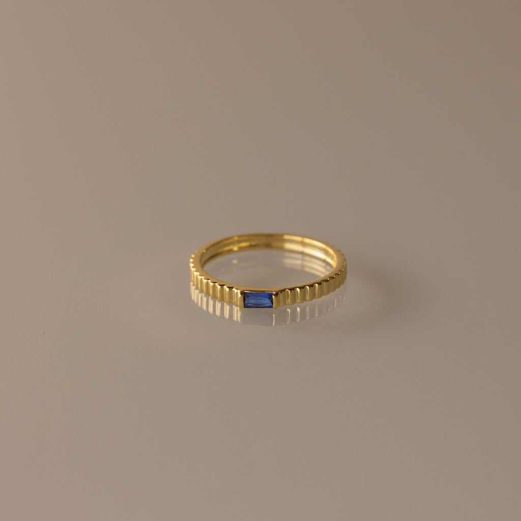 The Dainty Ribbed Sapphire Ring features a textured band and a petite rectangular blue sapphire, ideal for stacking and shown on a neutral background.