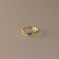 The Dainty Ribbed Sapphire Ring features a textured band and a petite rectangular blue sapphire, ideal for stacking and shown on a neutral background.