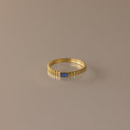 The Dainty Ribbed Sapphire Ring features a textured band and a petite rectangular blue sapphire, ideal for stacking and shown on a neutral background.