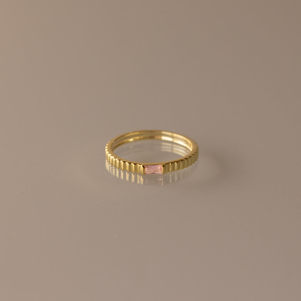 The Dainty Ribbed Pink Tourmaline Ring features a rectangular pink tourmaline gemstone at the center and a ribbed gold band, shown against a neutral background.
