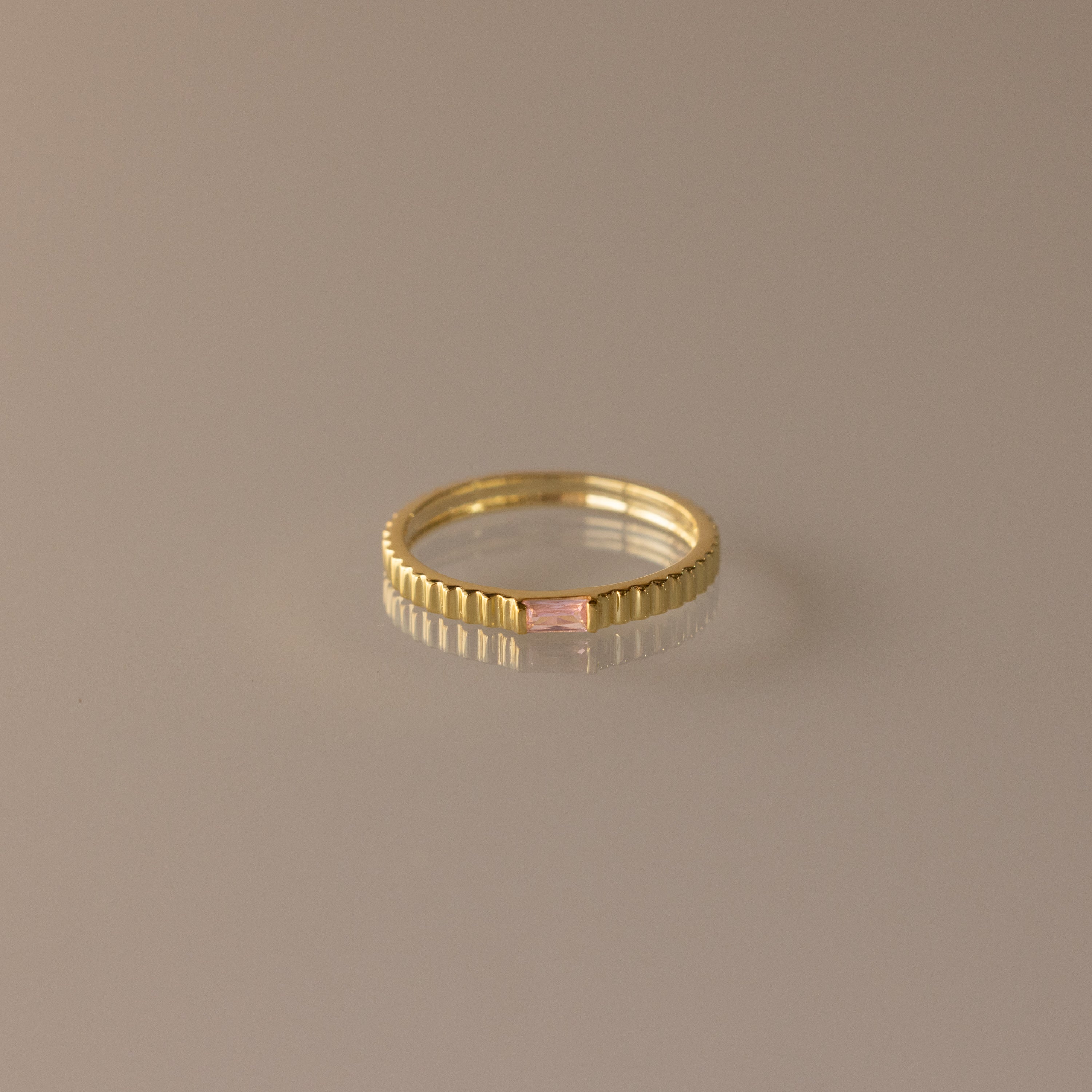 The Dainty Ribbed Pink Tourmaline Ring features a rectangular pink tourmaline gemstone at the center and a ribbed gold band, shown against a neutral background.