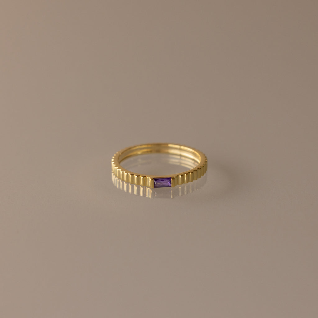 The Dainty Ribbed Amethyst Ring features a gold ribbed band and a small rectangular purple amethyst, perfect for stacking or as a delicate birthstone ring. Shown on a neutral background.