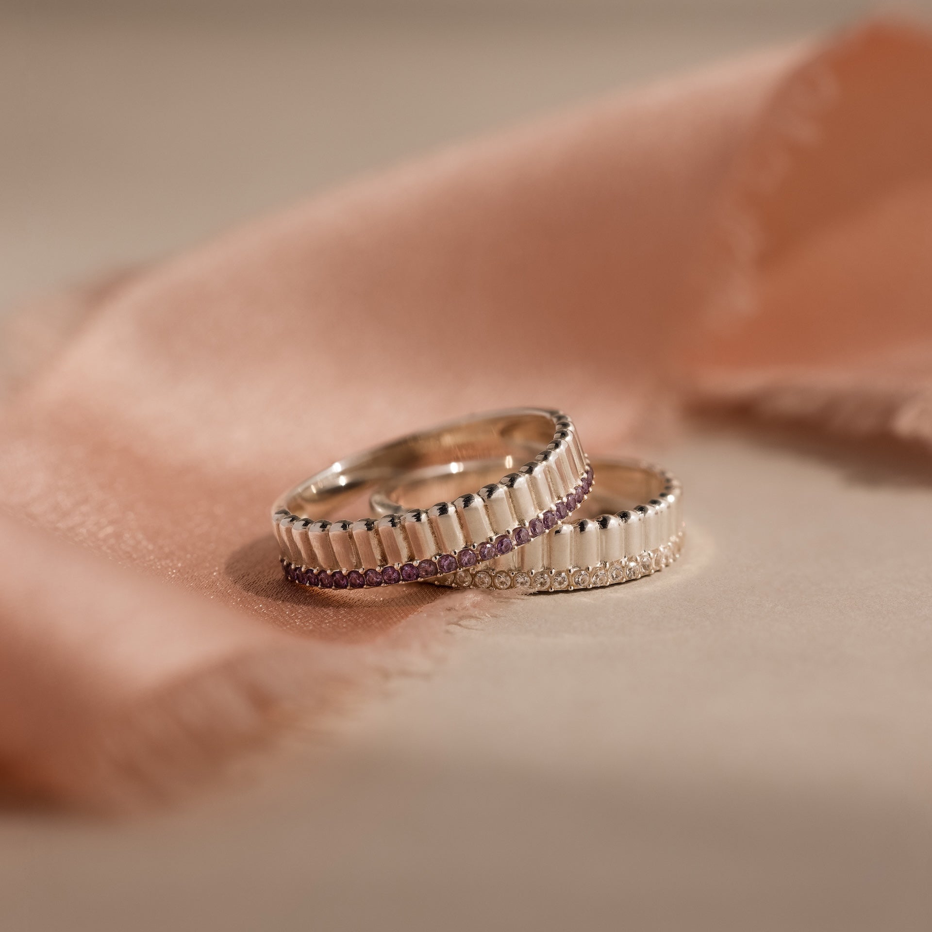 The Ribbed Birthstone Stack Ring, a silver ring with ribbed edges and small pink stones, is displayed on a soft peach fabric background.
