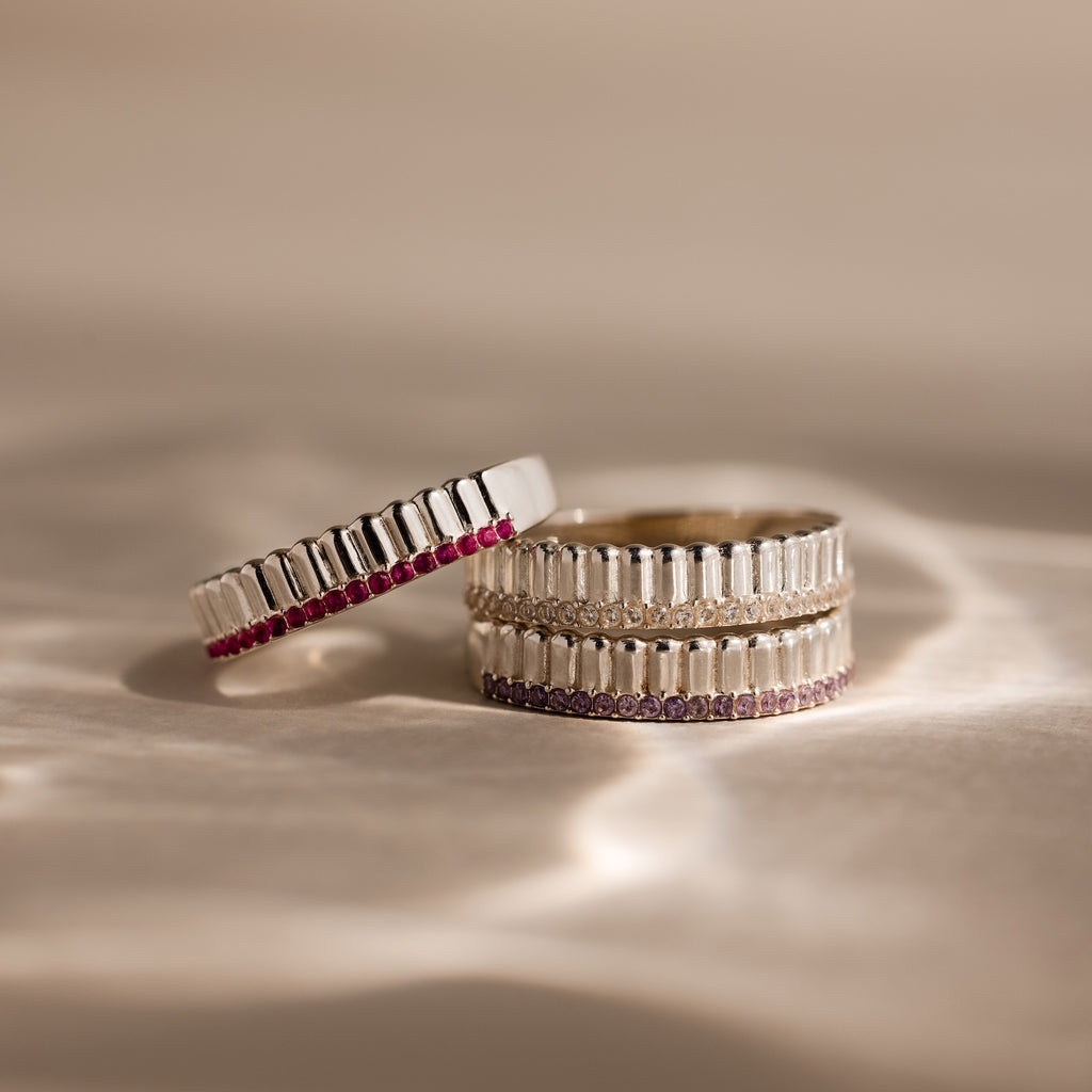 The Ribbed Garnet Stack Ring, featuring silver with baguette details and pink stones, lies on a textured beige surface—a perfect way to showcase your unique birthstone style.