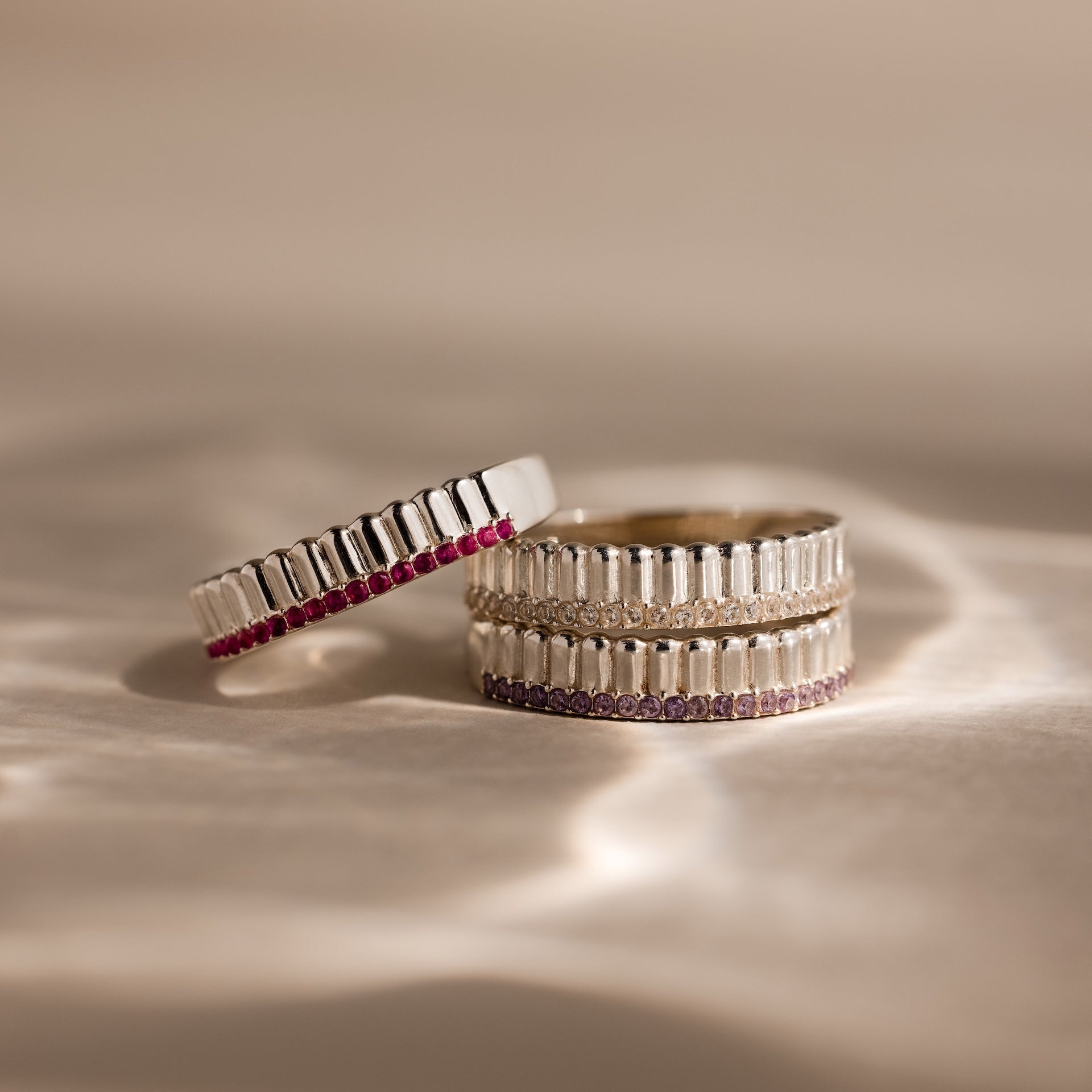 The Ribbed Garnet Stack Ring, featuring silver with baguette details and pink stones, lies on a textured beige surface—a perfect way to showcase your unique birthstone style.