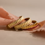 Three gold rings, including a Ribbed Garnet Stack Ring accented with pink, white, and red gemstones, rest elegantly on a soft pink fabric background.