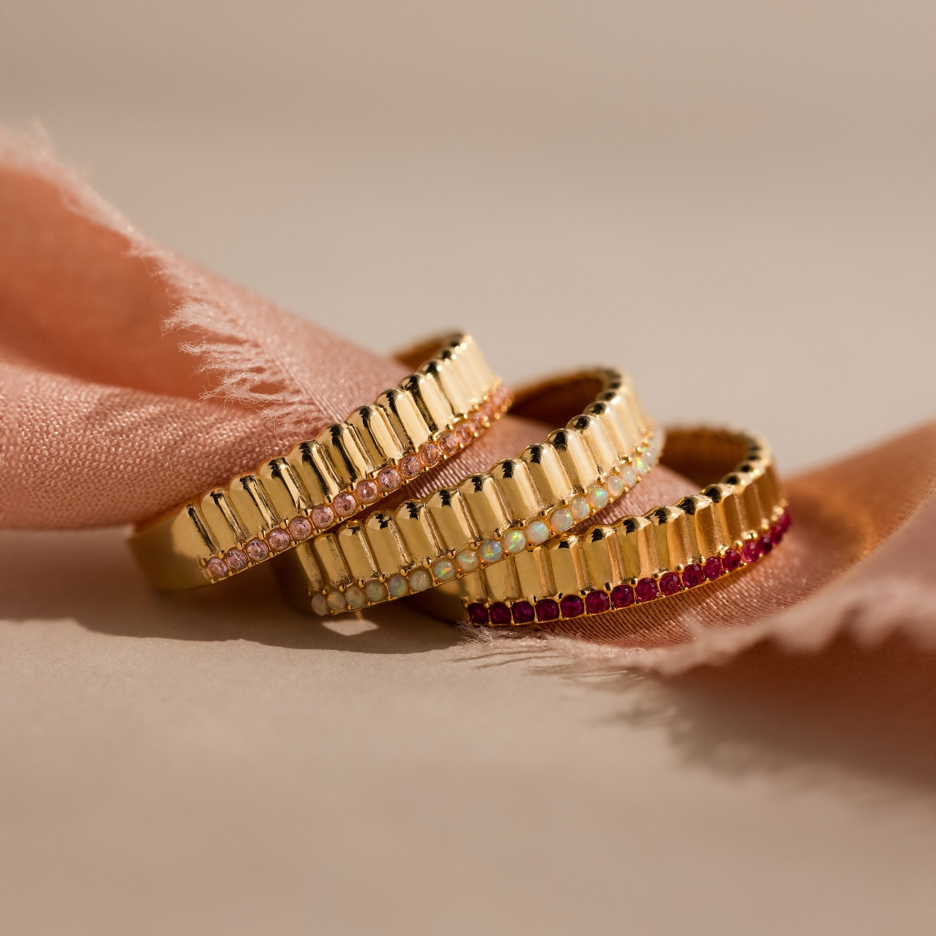 Three gold rings with colored gemstones, including a Ribbed Opal Stack Ring, displayed on a soft pink fabric background.