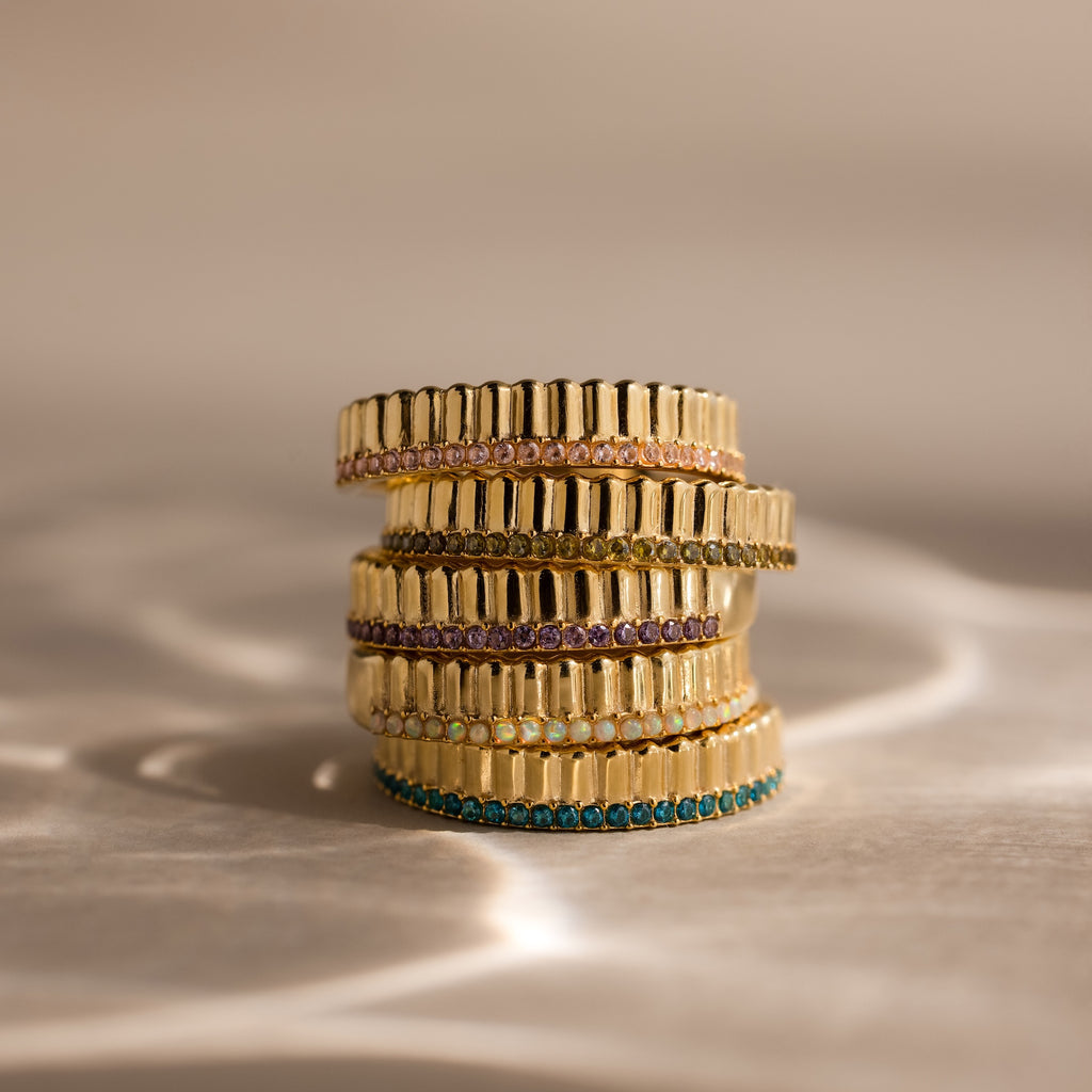 The Ribbed Blue Zircon Stack Ring features stacked gold bands set with vibrant blue zircon gemstones, shining beautifully on a reflective surface.