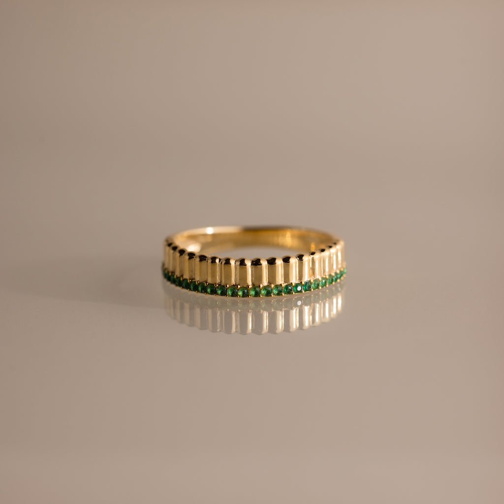 The Ribbed Emerald Stack Ring is a gold band with a sculpted ribbed texture and a row of small green emeralds at the center, perfect for everyday ring stacks and shown against a plain background.
