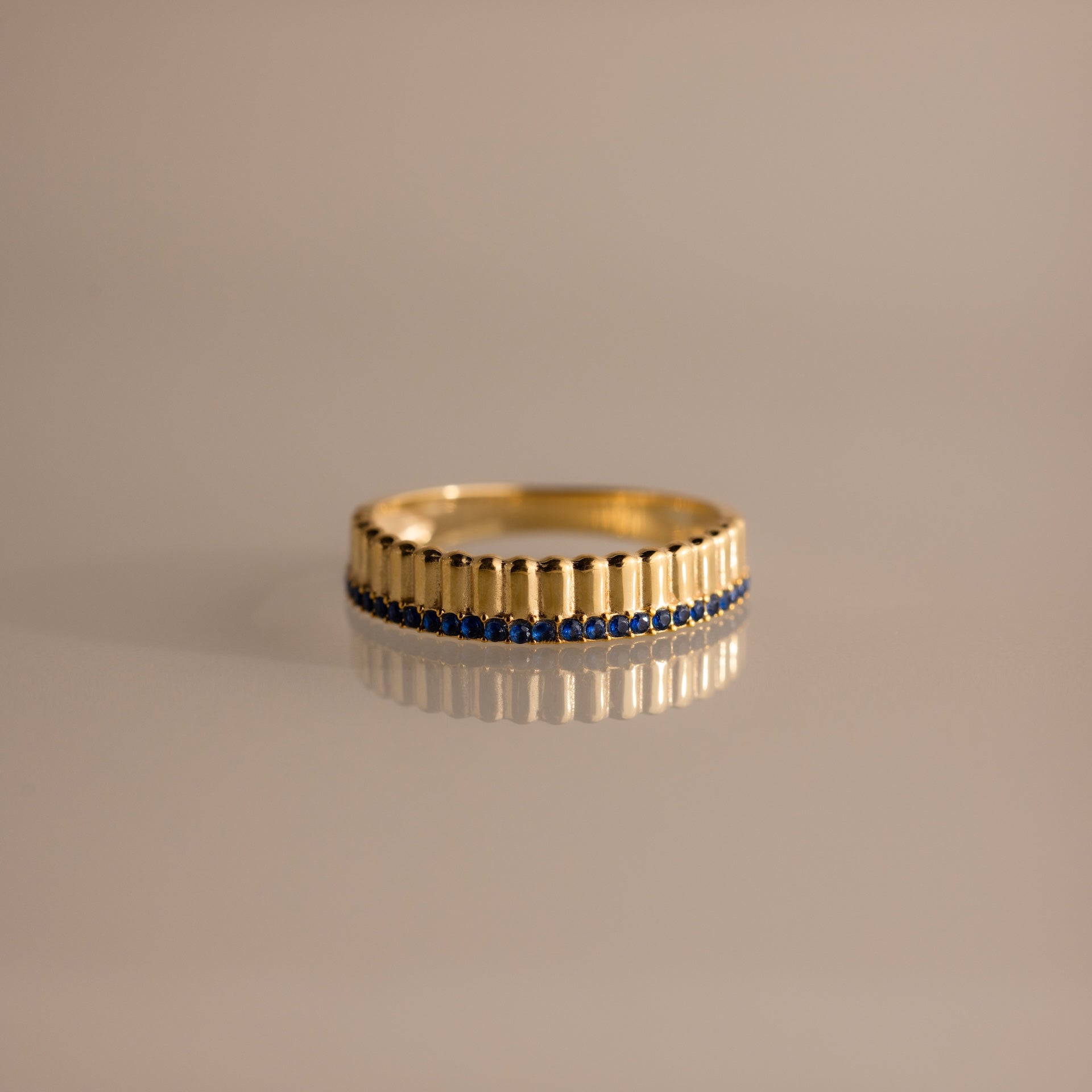 The Ribbed Sapphire Stack Ring features a sculpted ribbed texture, vertical engravings, and a row of small blue sapphires, shown on a reflective surface.