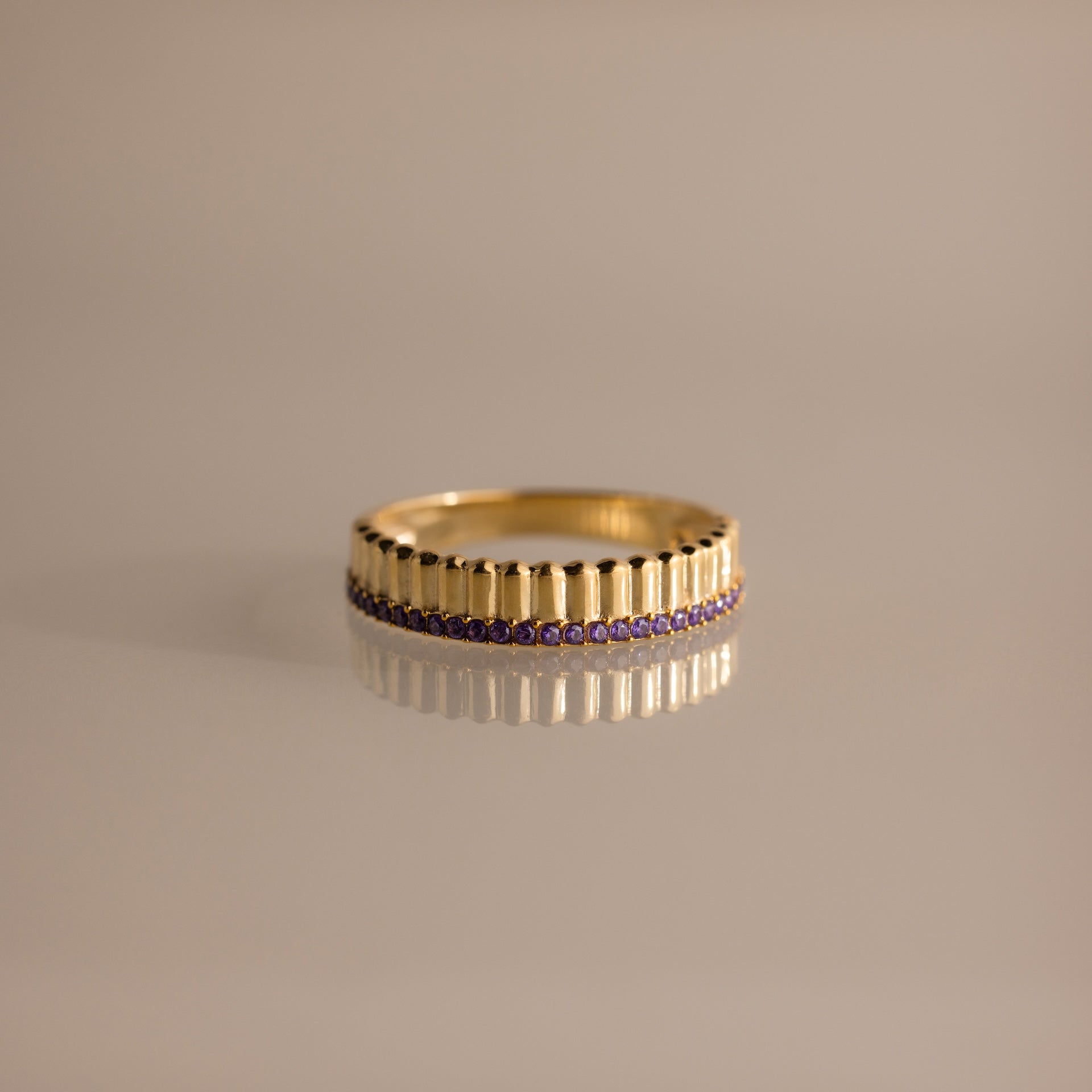 The Ribbed Amethyst Stack Ring features gold ribbed texture, vertical grooves, and a row of small purple amethyst stones. Reflected on a beige surface, it's perfect for stacking and adding elegant birthstone color to your collection.