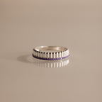 The Ribbed Amethyst Stack Ring, featuring a silver ribbed texture and a row of small purple stones, is shown on a reflective beige surface. This stylish birthstone ring is perfect for stacking and layering.