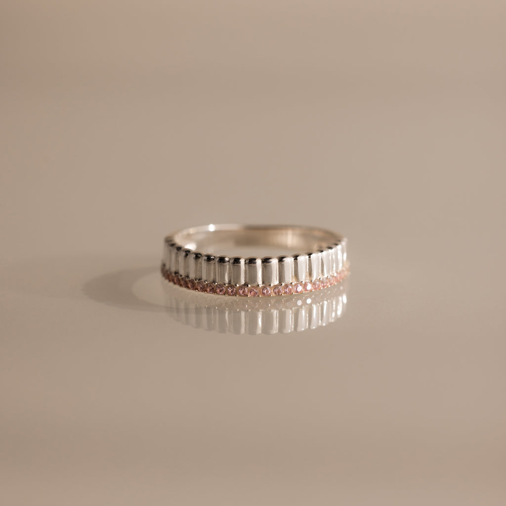 The Ribbed Pink Tourmaline Stack Ring features a sculpted ribbed texture and a row of small pink tourmaline gemstones, shown on a reflective surface. Ideal for creating your own unique ring stack style.