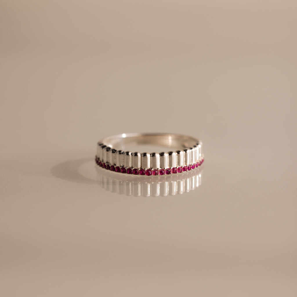 Ribbed Ruby Stack Ring with a row of small red gemstones set in the center, shown on a beige surface.