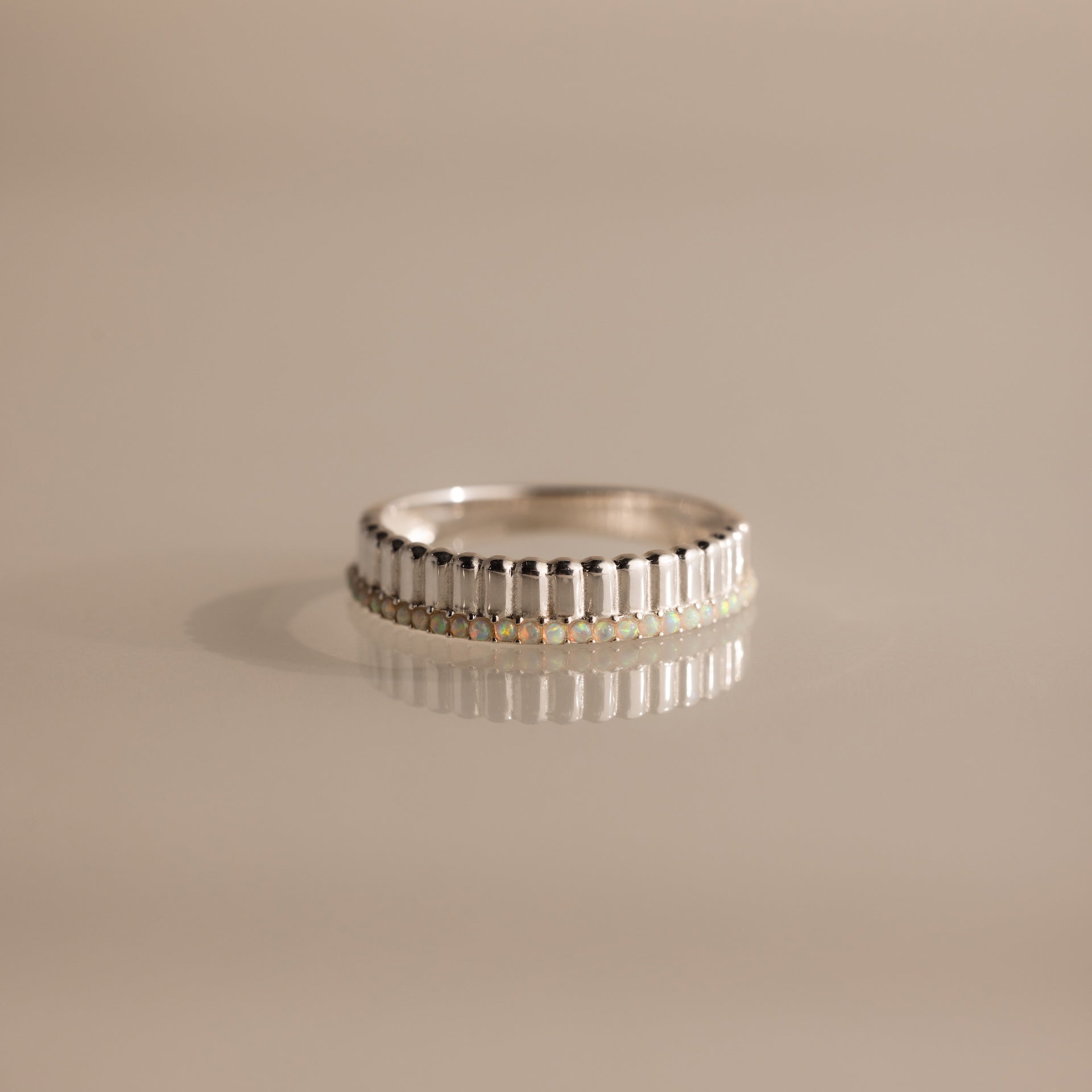 The Ribbed Opal Stack Ring features a row of small opal stones set in a silver ribbed band, shown on a beige surface—ideal for enhancing your favorite ring stack.