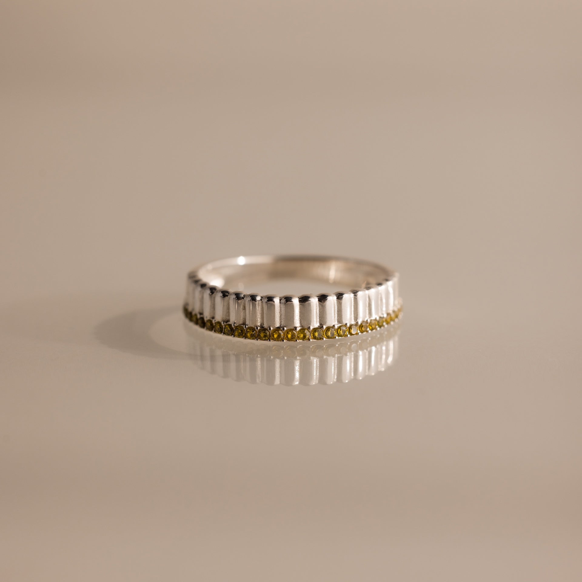 The Ribbed Peridot Stack Ring features a silver ribbed texture with vertical grooves and a row of small yellow peridot stones, shown on a neutral background. A modern, stackable ring perfect for versatile styling.