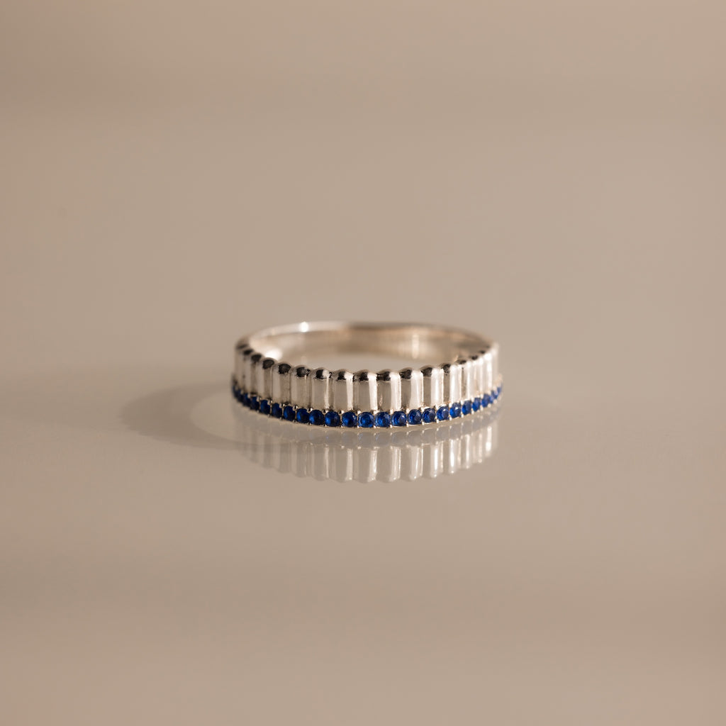 The Ribbed Sapphire Stack Ring is a modern piece with sculpted ribbed texture, vertical ridges, and a row of small blue sapphires set around the band, displayed on a beige background.