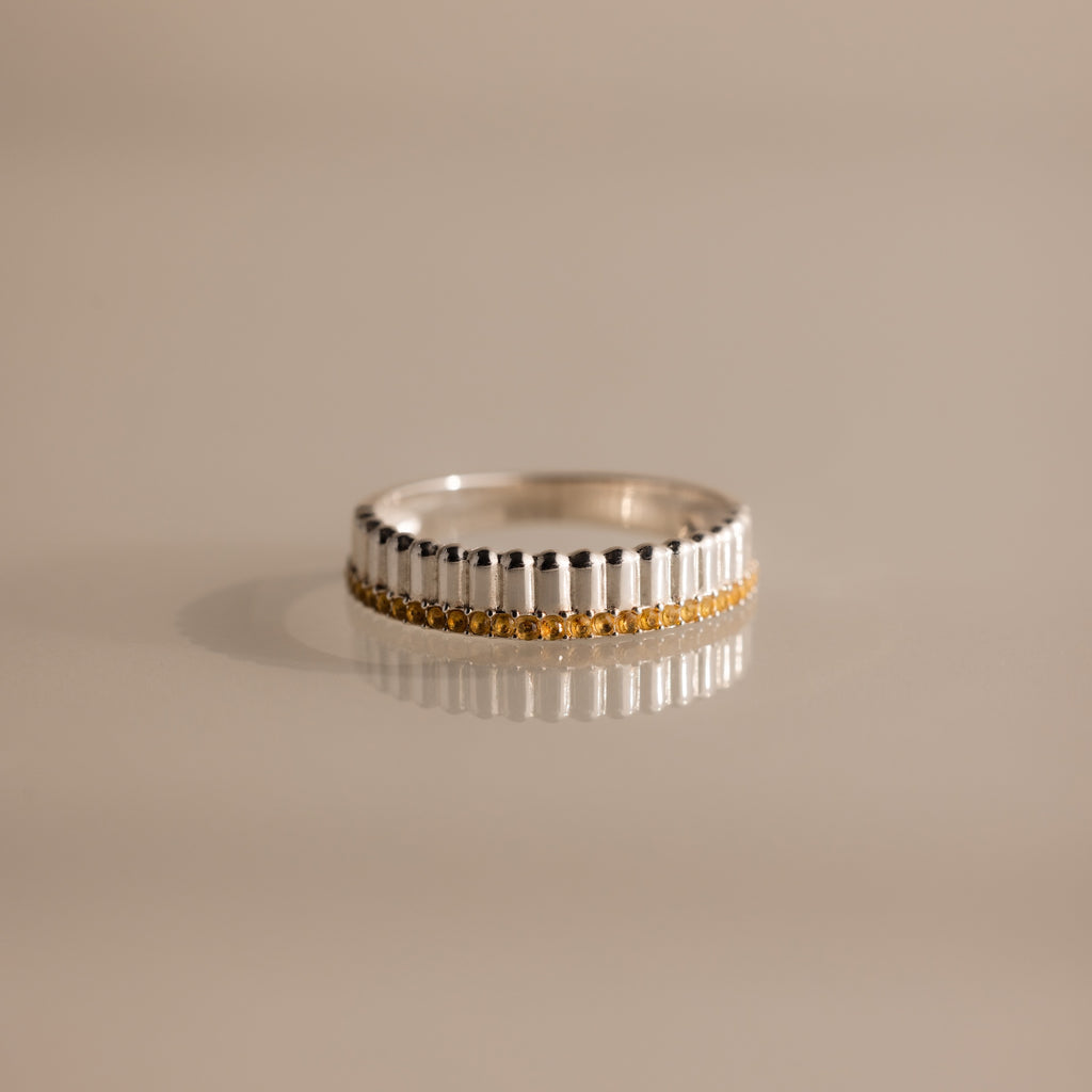 The Ribbed Topaz Stack Ring features a sculpted ribbed texture and a row of small yellow topaz stones set around the band, displayed on a shiny surface.