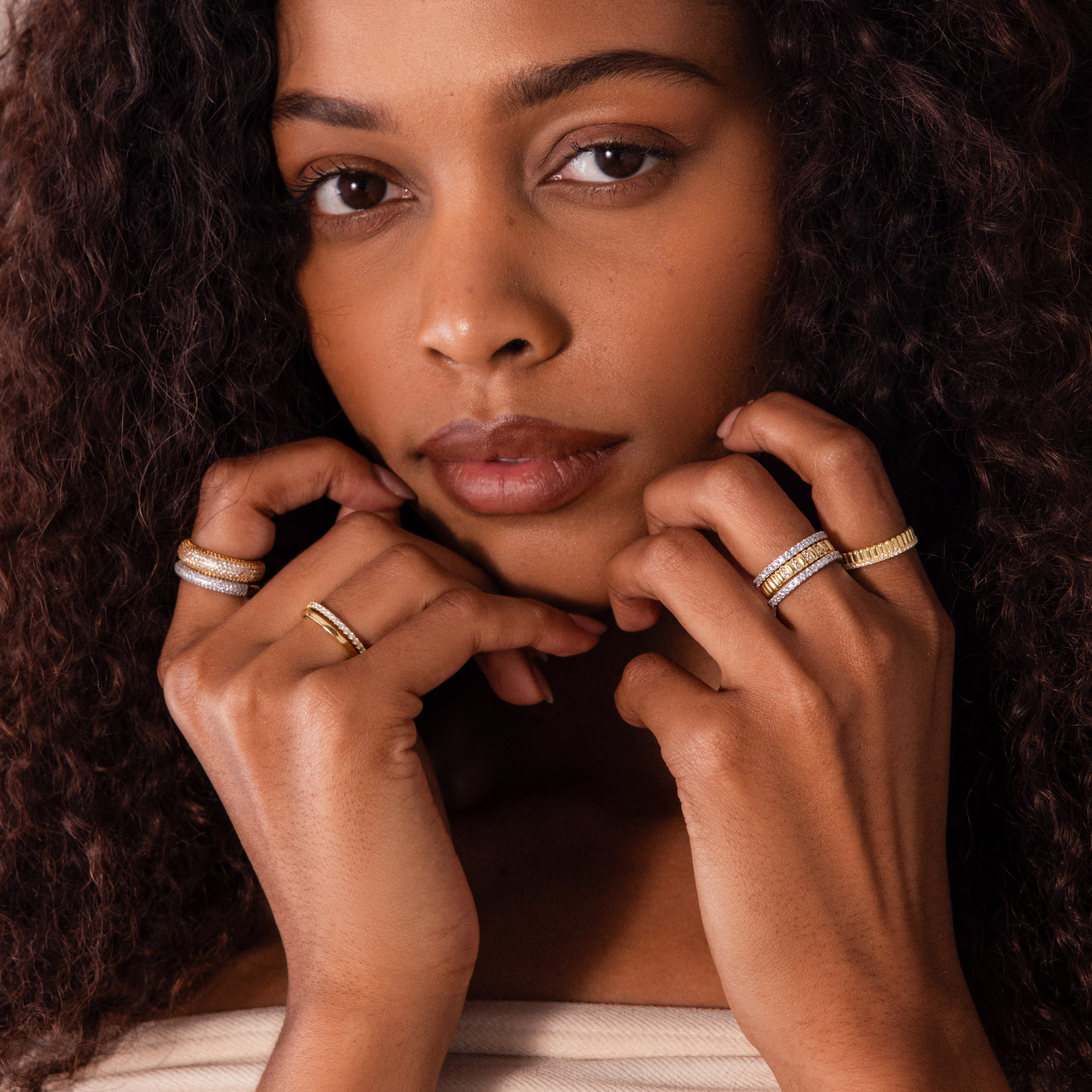 Woman with curly hair wearing multiple gold and silver rings, including a Ribbed Diamond Stack Ring, gently touching her face with both hands.