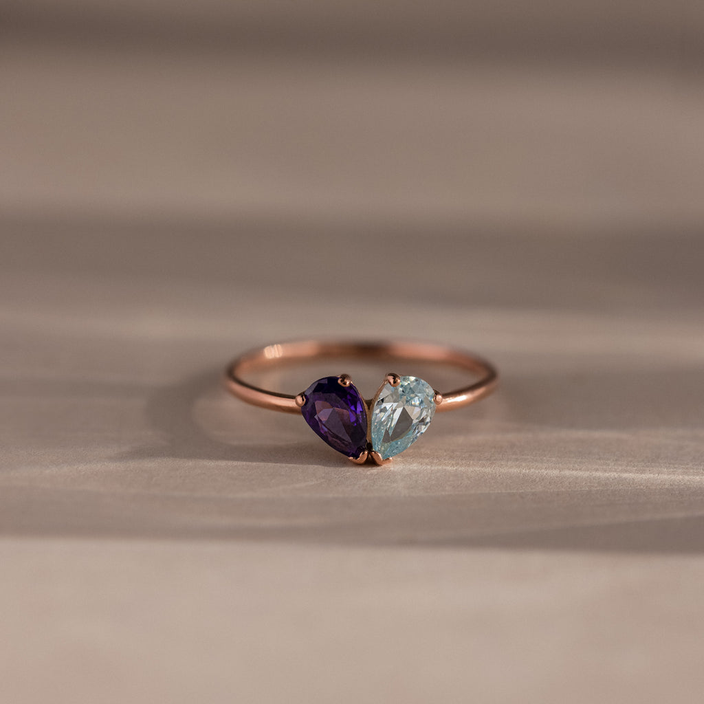 The Toi et Moi Teardrop Heart Ring in Rose Gold showcases heart-shaped purple and light blue gemstones set side by side, displayed on a neutral background.