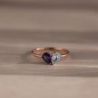 The Toi et Moi Teardrop Heart Ring in Rose Gold showcases heart-shaped purple and light blue gemstones set side by side, displayed on a neutral background.