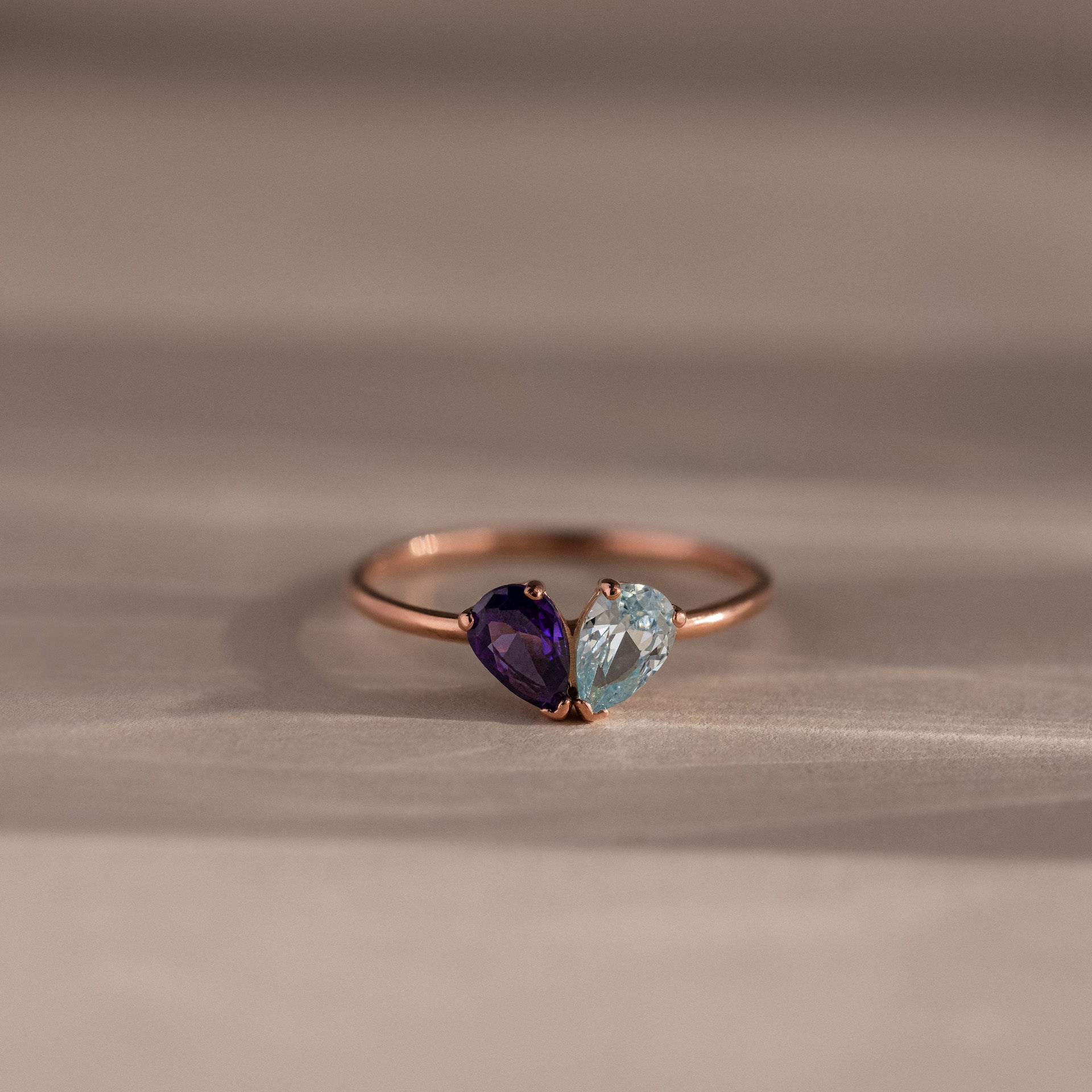The Toi et Moi Teardrop Heart Ring in Rose Gold showcases heart-shaped purple and light blue gemstones set side by side, displayed on a neutral background.