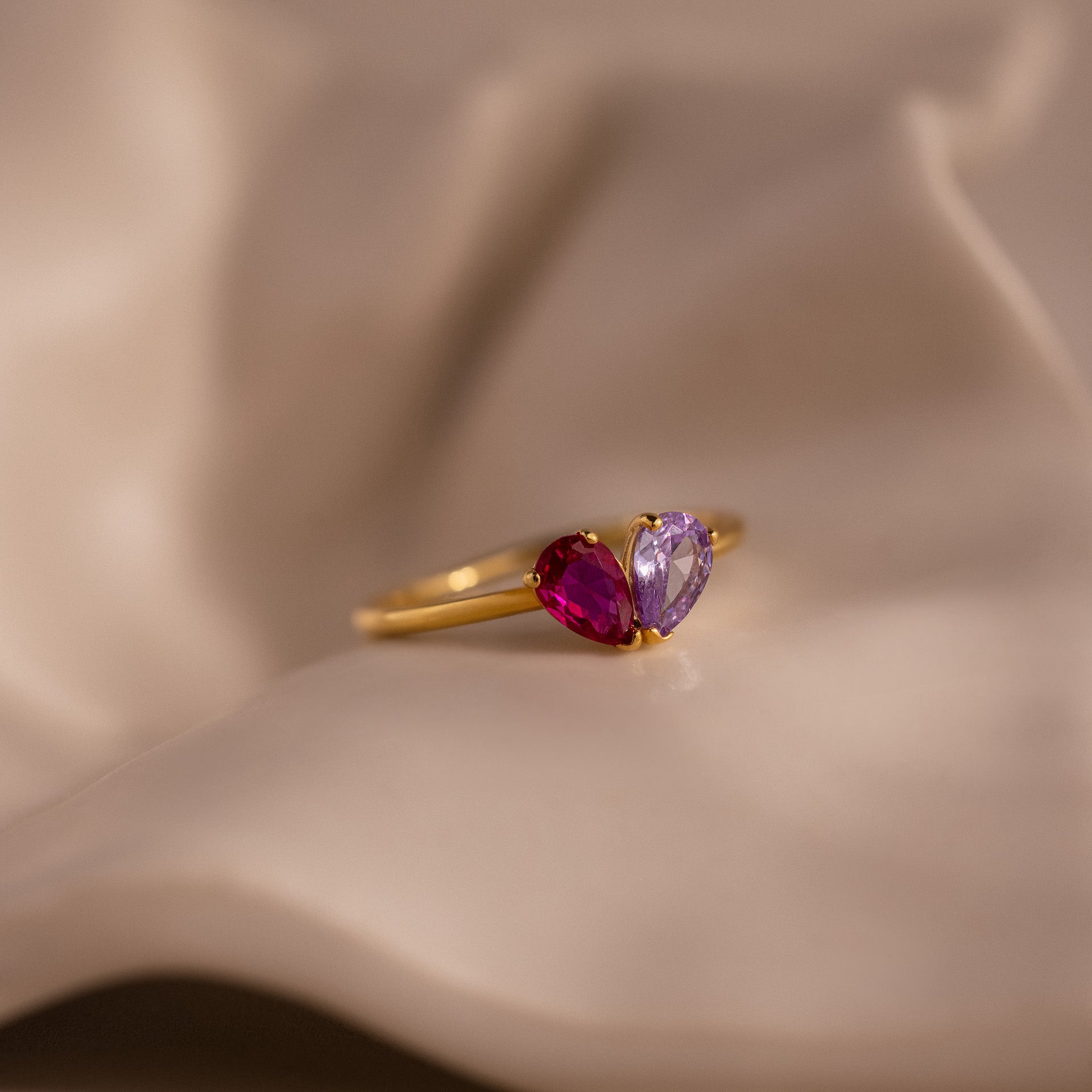 The Toi et Moi Teardrop Heart Ring in gold, featuring one pink and one purple heart-shaped gemstone, rests elegantly on a soft beige surface.