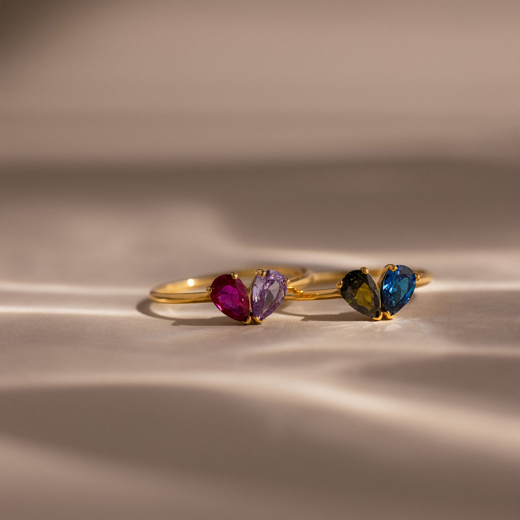 Two Toi et Moi Teardrop Heart Rings, each with two heart-shaped gemstones in vivid colors, are elegantly displayed on a soft beige surface.