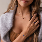 A woman in a brown top and grey wrap wears gold jewelry with blue stones, including the Toi et Moi Teardrop Heart Ring, a pear-cut stone necklace, and a matching bracelet.