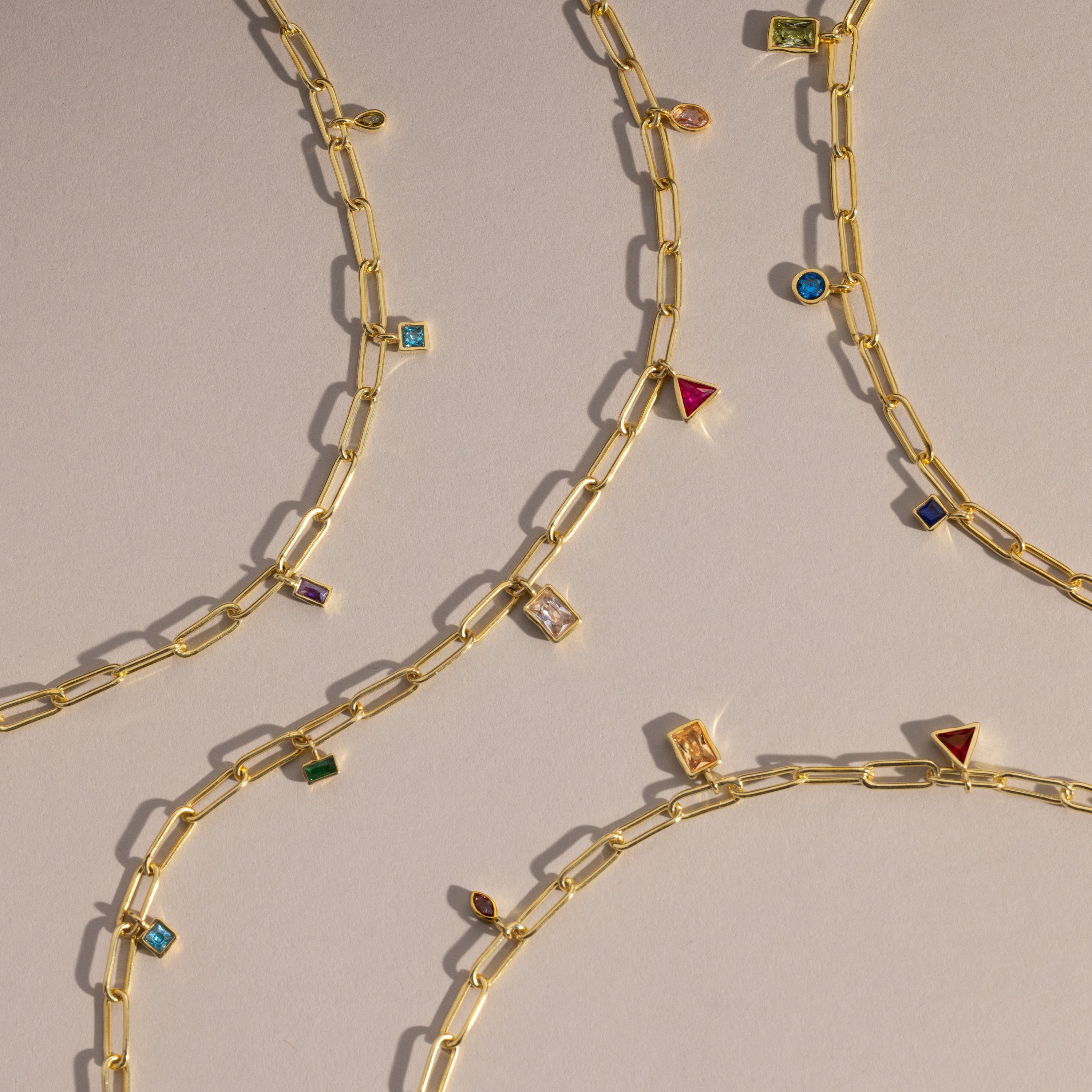 The Mixed Birthstone Charm Necklace in 18K Gold is displayed diagonally on a light beige surface, adding a touch of unique jewelry style with its colorful gemstone charms.