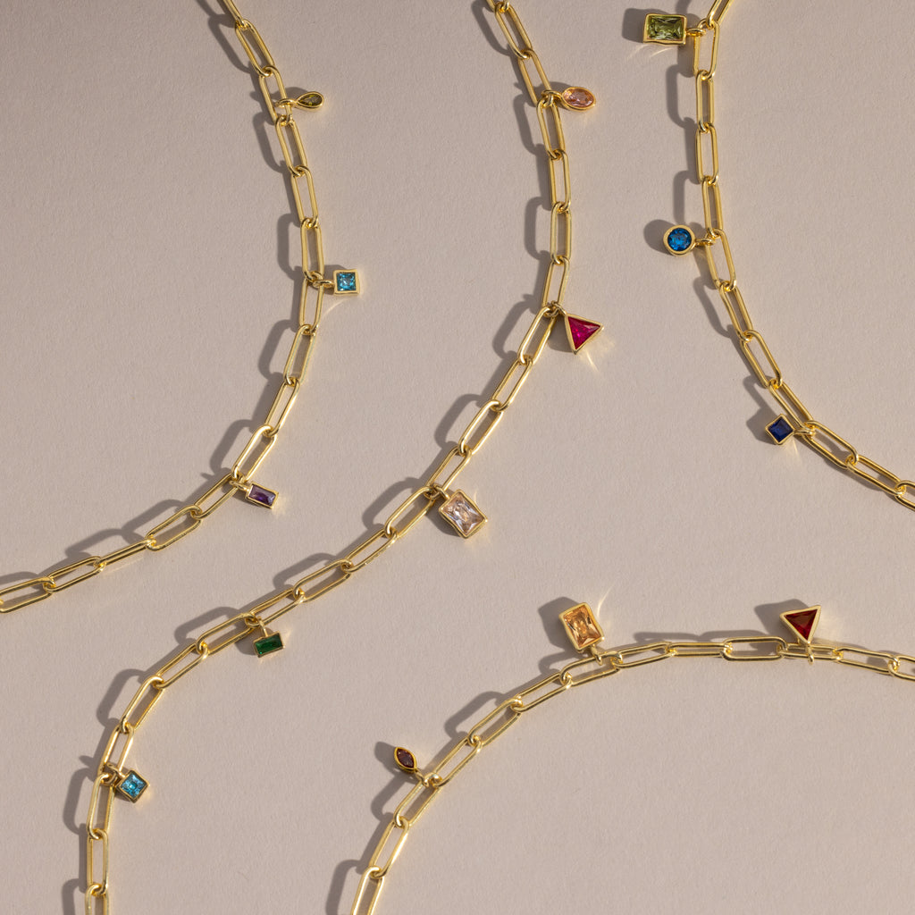 The Mixed Birthstone Charm Necklace features gold chains with unique charms and colorful gemstone accents, arranged diagonally on a neutral background—ideal for personalized jewelry lovers.
