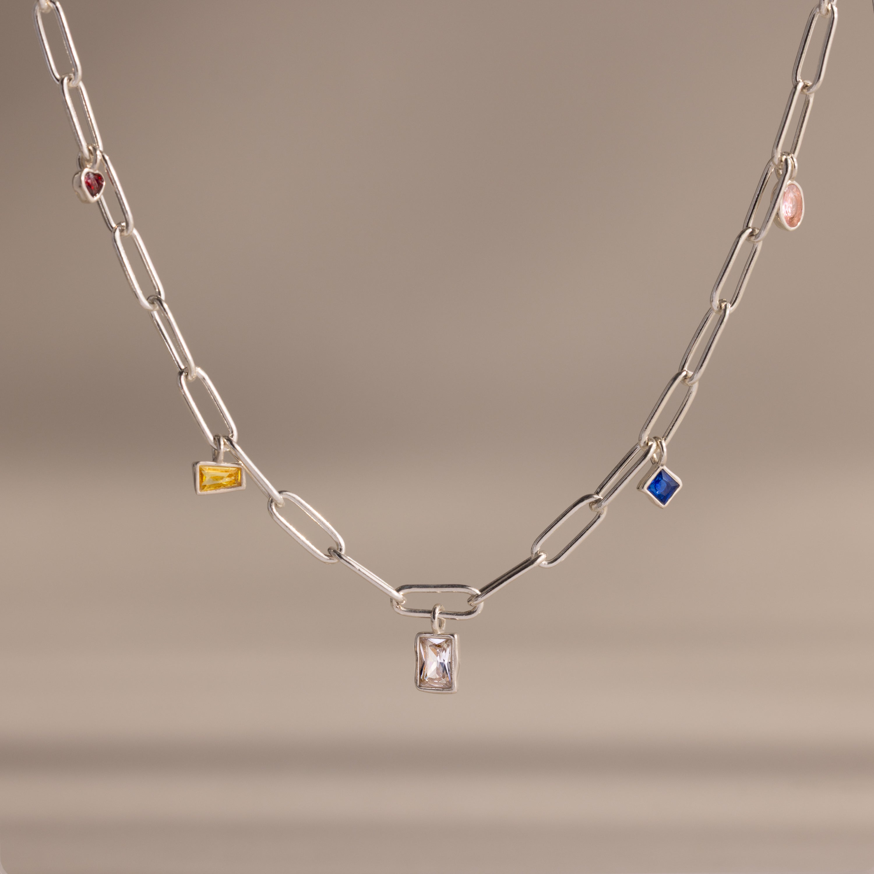 The Mixed Birthstone Charm Necklace in Sterling Silver features colorful gem charms in yellow, blue, pink, and red, displayed on a silver chain against a beige background. This unique piece adds vibrant style to any outfit.
