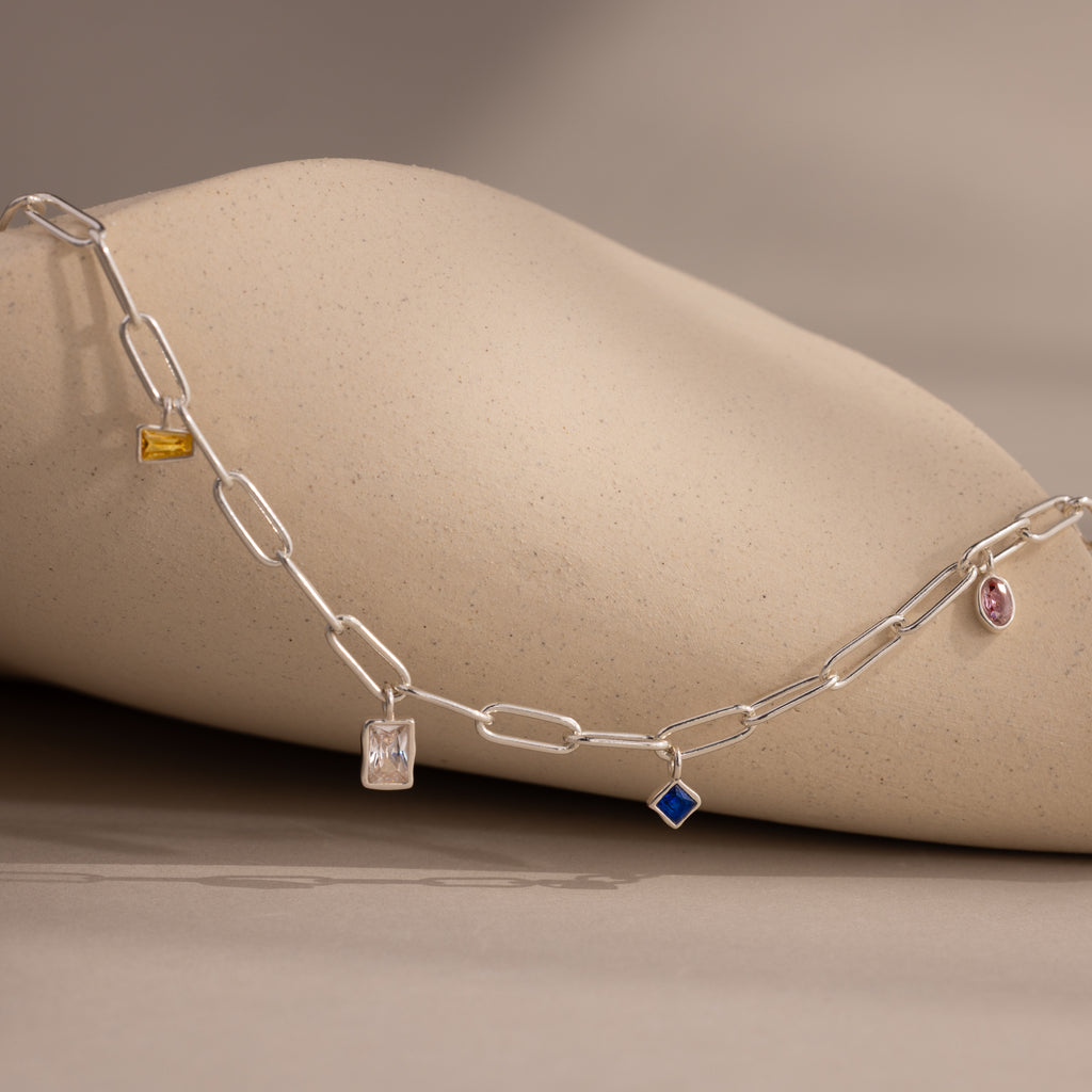 The Mixed Birthstone Charm Necklace, featuring distinctive charms, rests gracefully against a soft beige stone background.