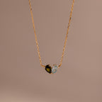 The Toi et Moi Teardrop Heart Necklace features a gold chain and a heart pendant with half black, half clear gemstones, shown on a beige background.