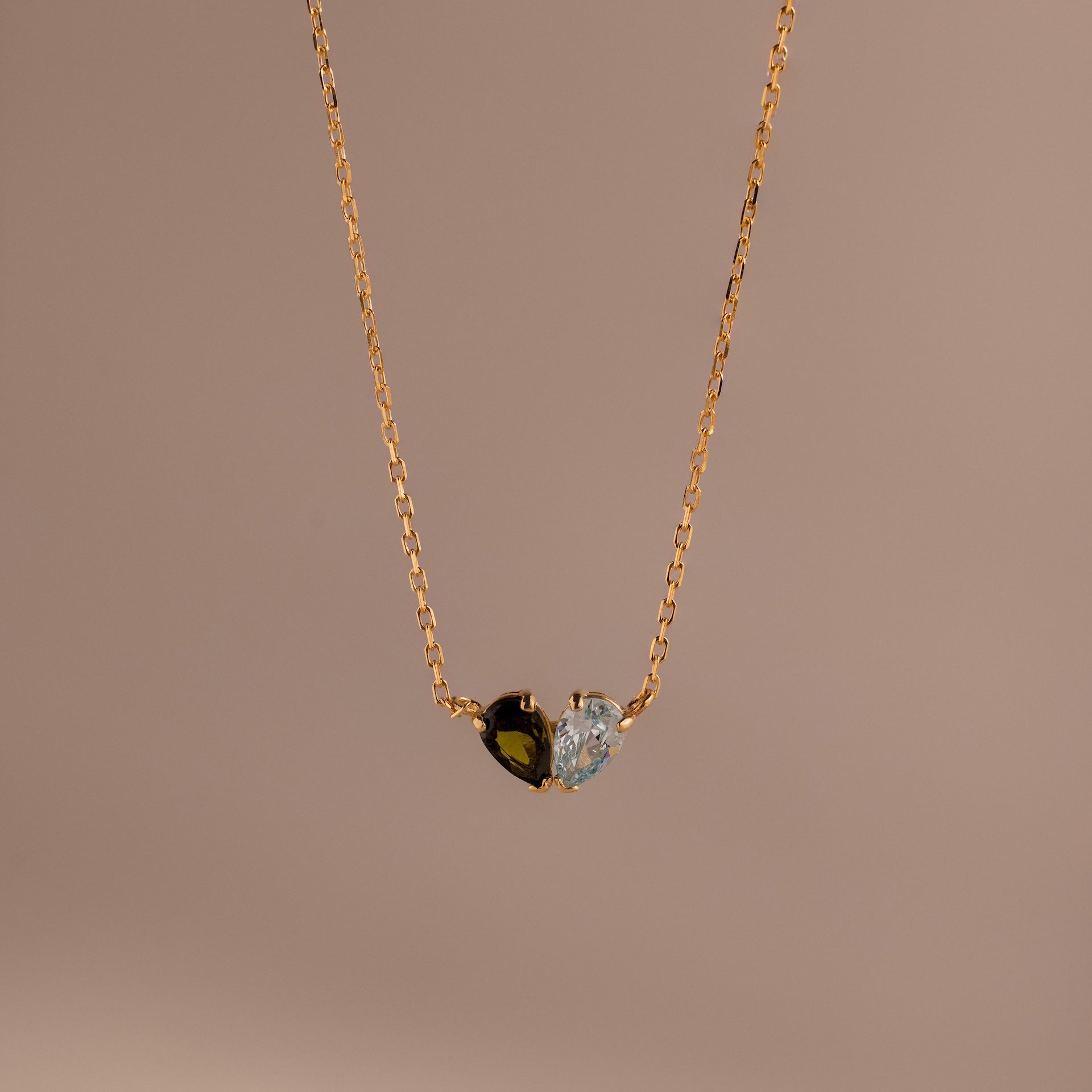 The Toi et Moi Teardrop Heart Necklace features a gold chain and a heart pendant with half black, half clear gemstones, shown on a beige background.