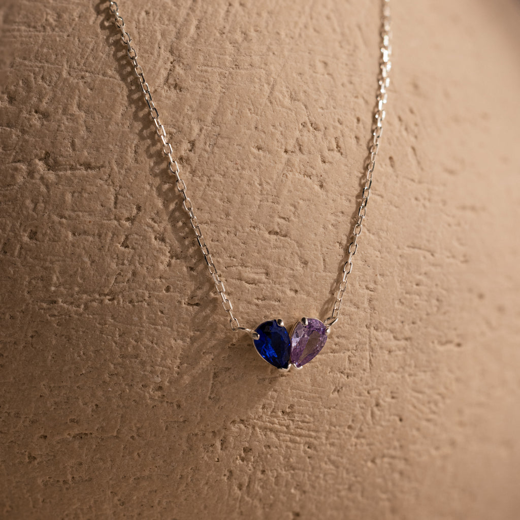 The Toi et Moi Teardrop Heart Necklace showcases pear-cut blue and purple gemstones, elegantly displayed on a textured beige surface.