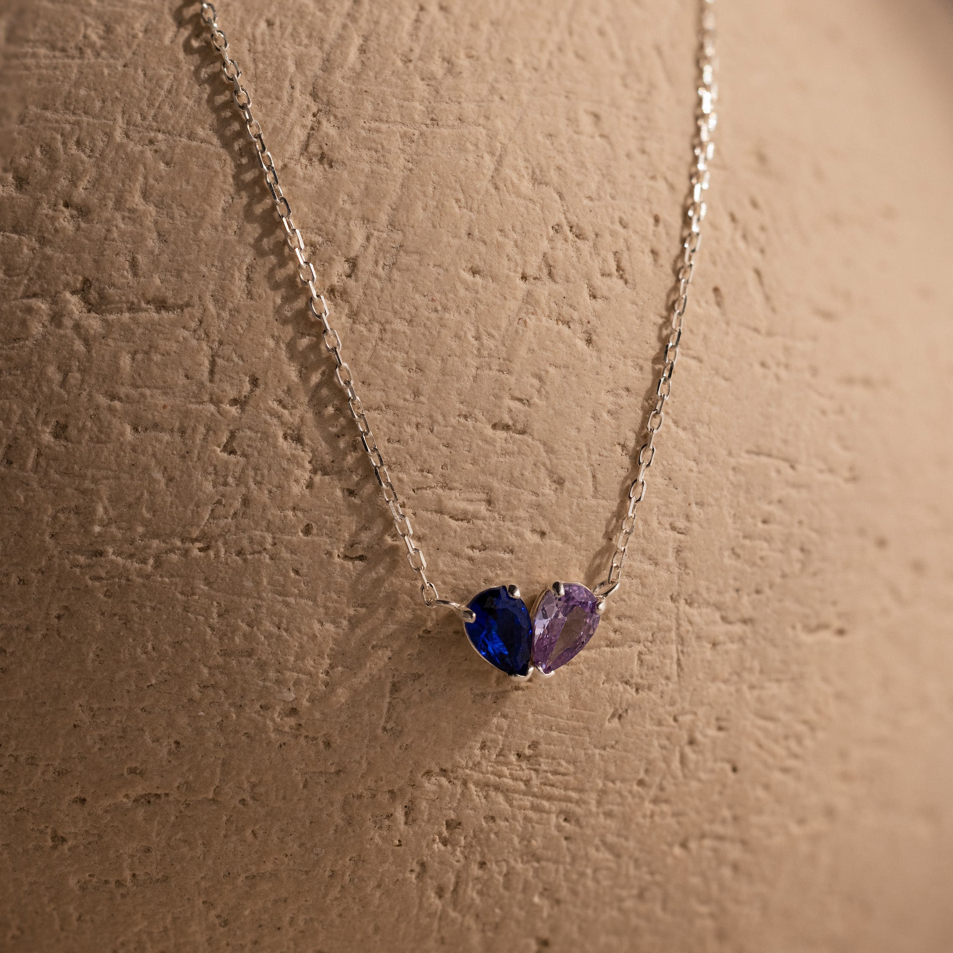 The Toi et Moi Teardrop Heart Necklace showcases pear-cut blue and purple gemstones, elegantly displayed on a textured beige surface.