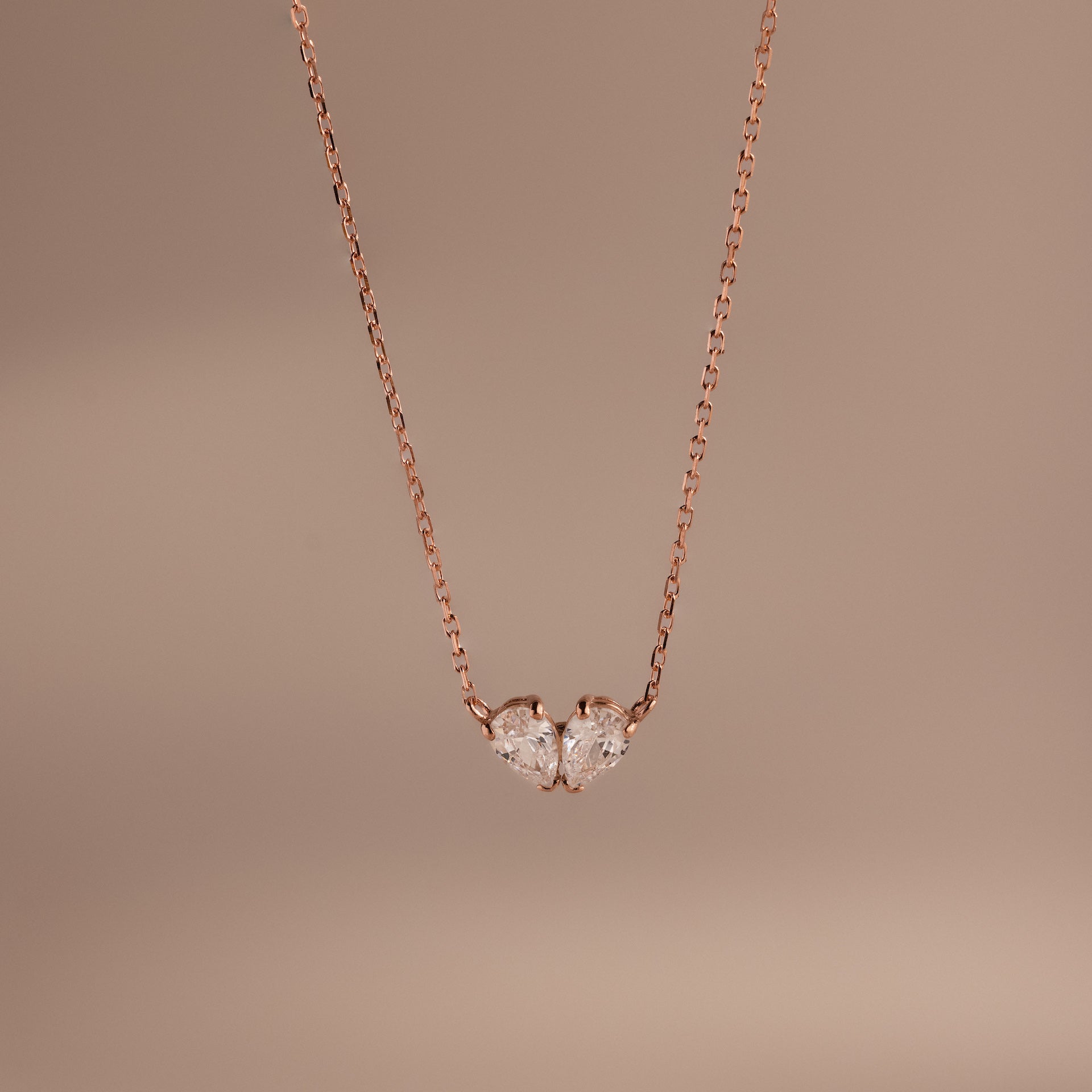 Toi et Moi Teardrop Heart Necklace in Rose Gold features two heart-shaped clear gemstones against a simple, soft beige background.
