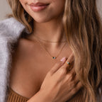 A woman in a brown top touches her chest while wearing the Toi et Moi Teardrop Heart Necklace in 18K Gold and a gold ring with blue stones.