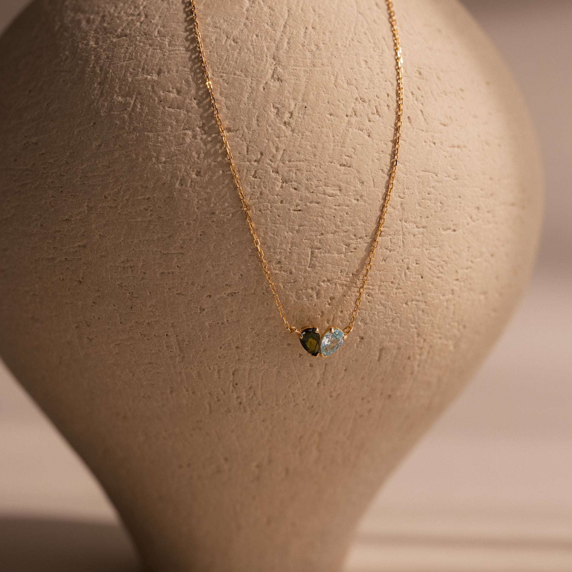 The Toi et Moi Teardrop Heart Necklace in 18K Gold, featuring two small heart-shaped pendants, is displayed against a textured beige vase background.